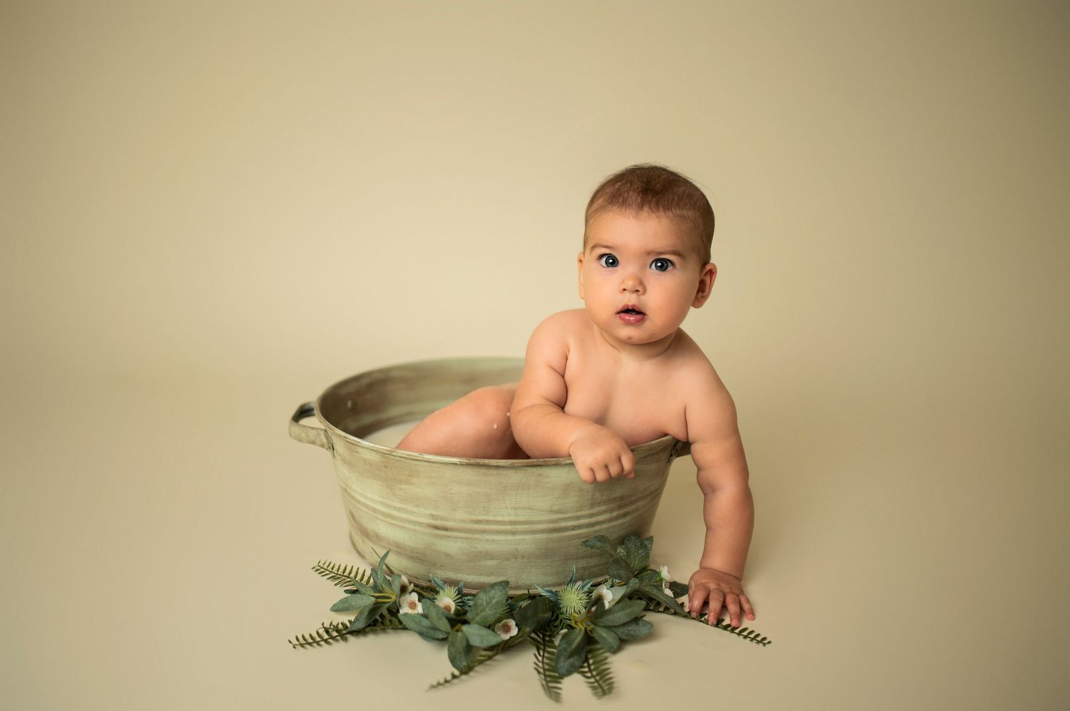 Baby in antique wash tub decorated with greenery against beige background.