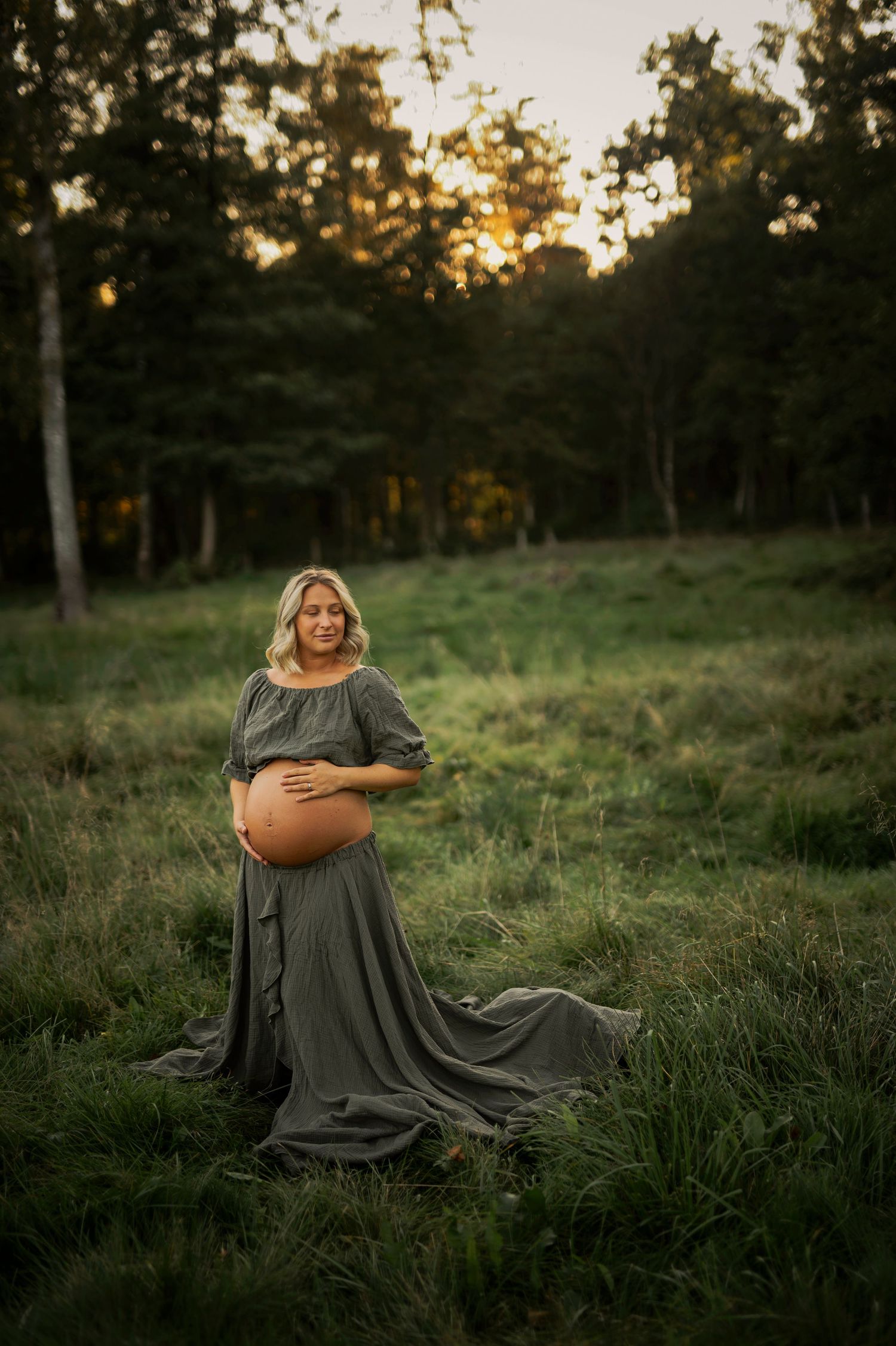 Maternity photo of a woman in gray dress standing in grassy field during sunset.
