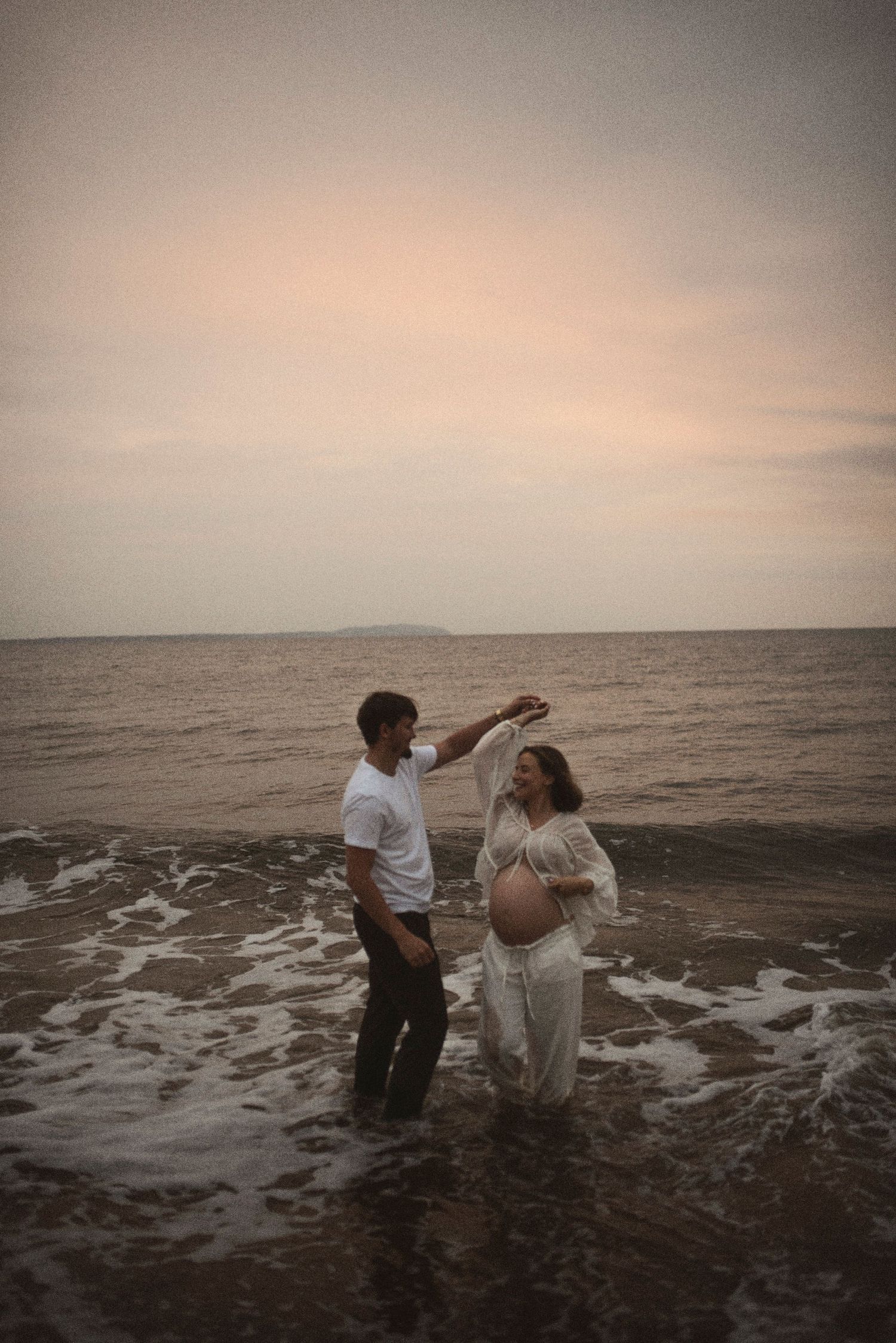 Expectant couple shares a tender moment while standing in gentle ocean waves at sunset.