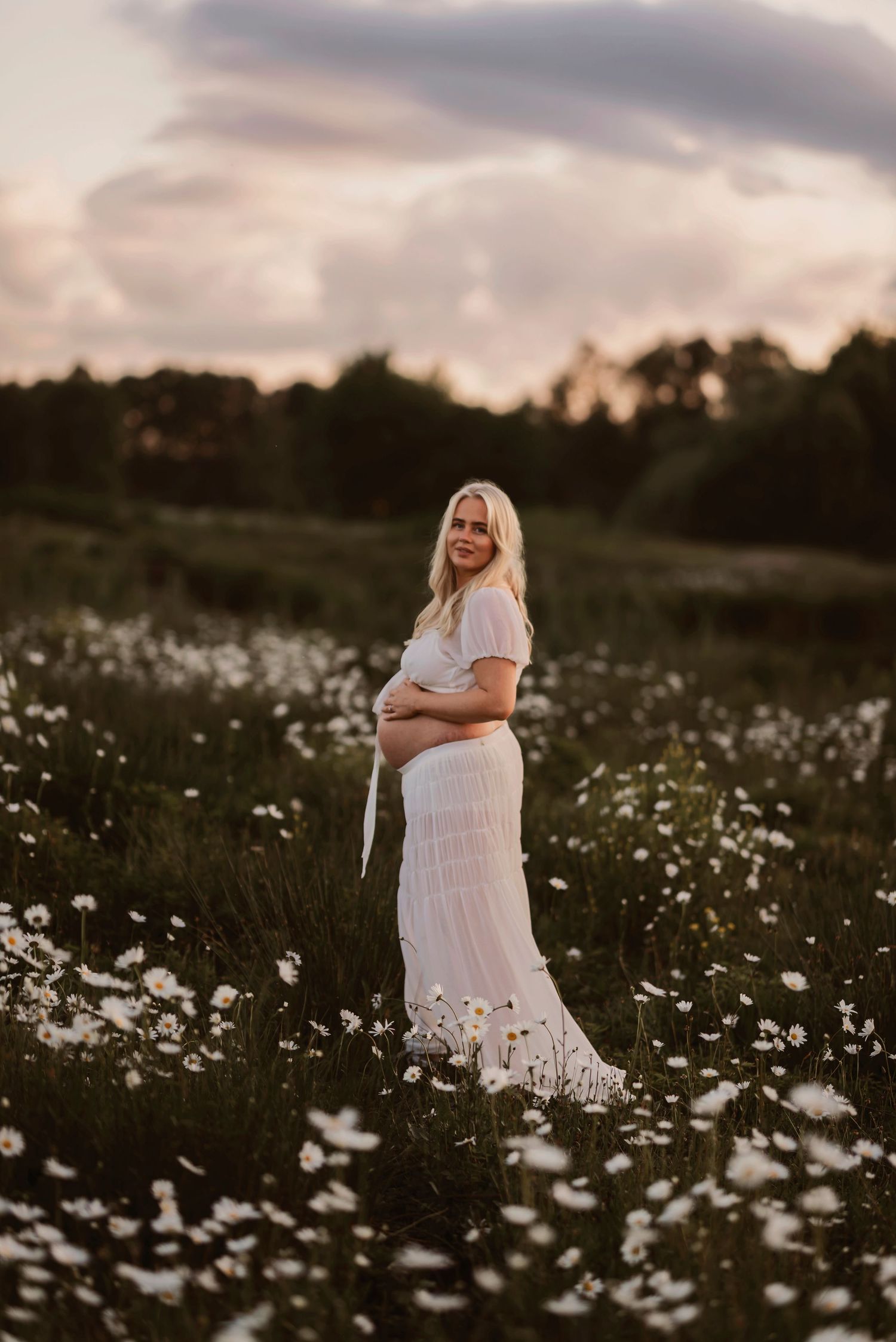 Expectant mother in flowing white dress poses gracefully among blooming daisies at dusk.