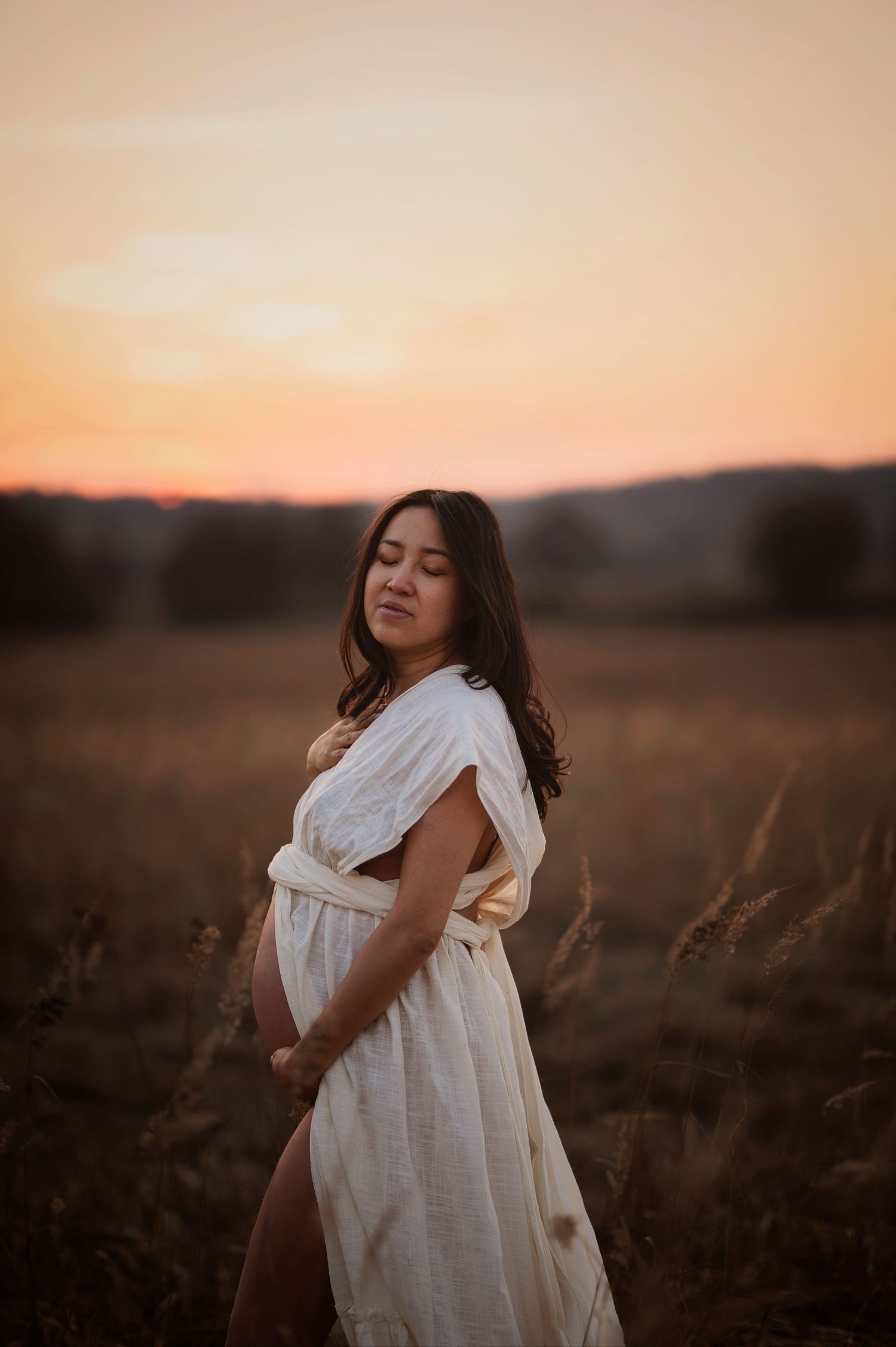 A pregnant woman in a flowing white dress stands in a field at sunset with golden light illuminating the scene.