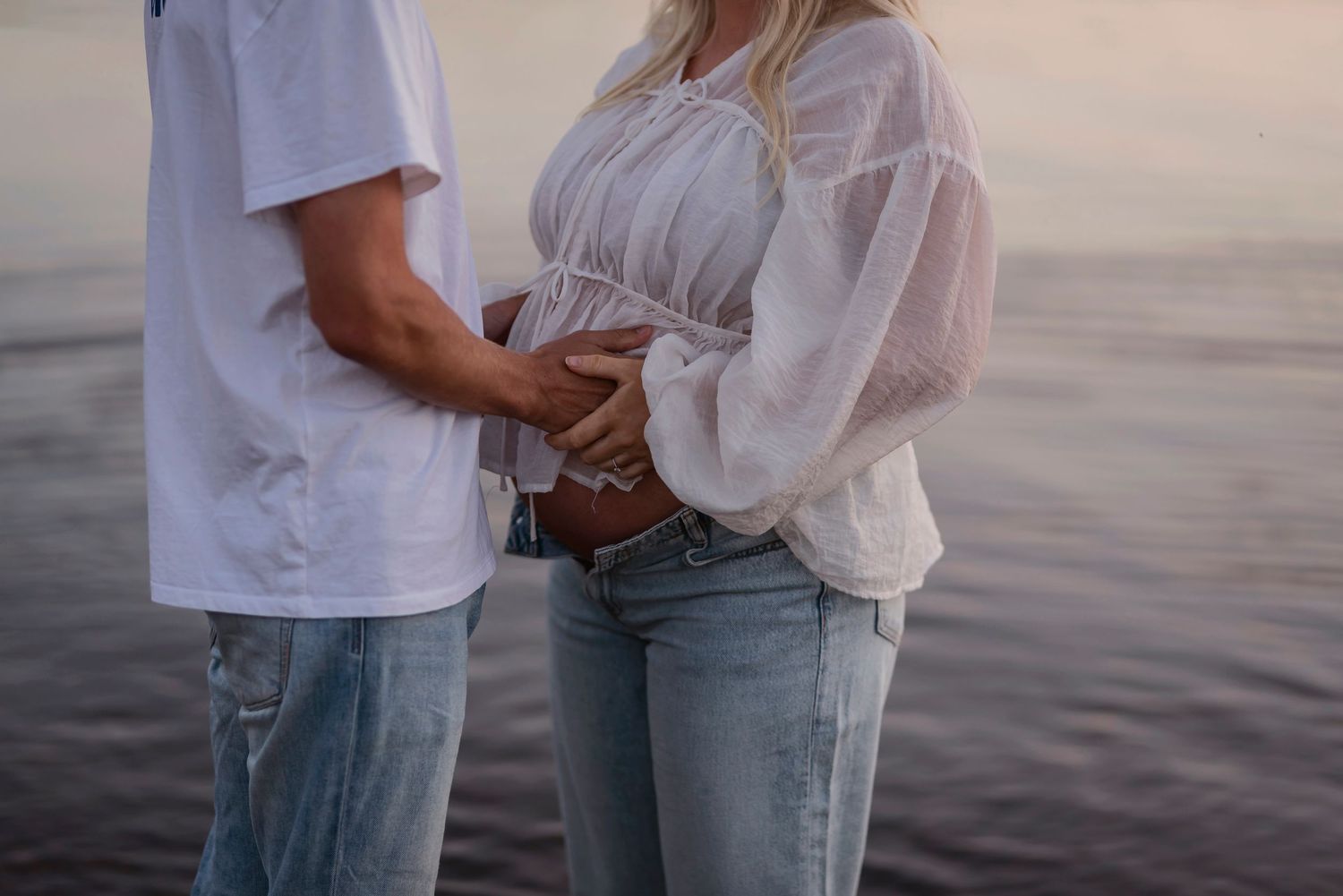 Couple in casual wear embracing by a serene lake during sunset.