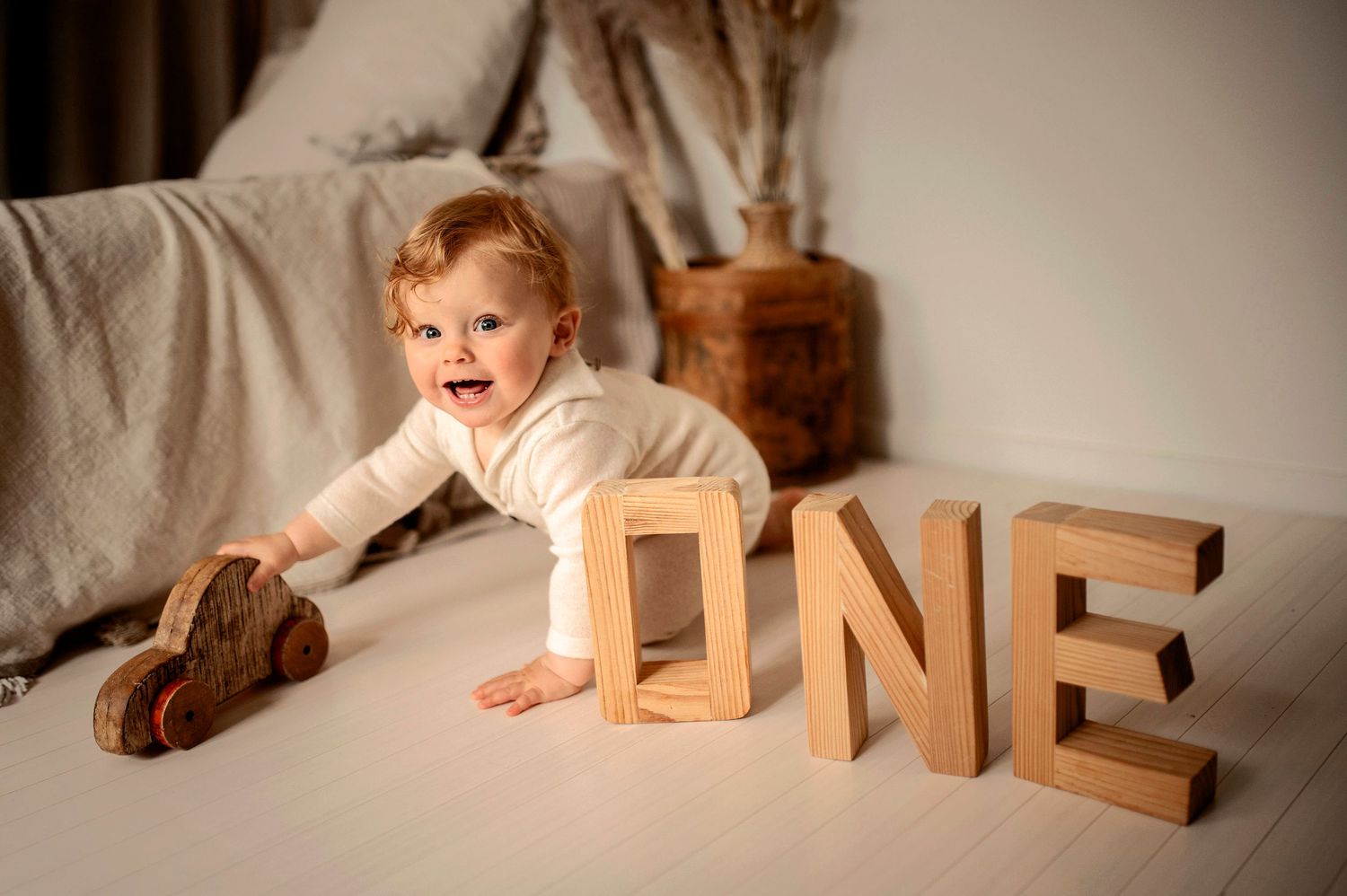 Baby in white outfit crawls on couch next to large wooden 'ONE' letters and stuffed animal toy in cozy home setting.