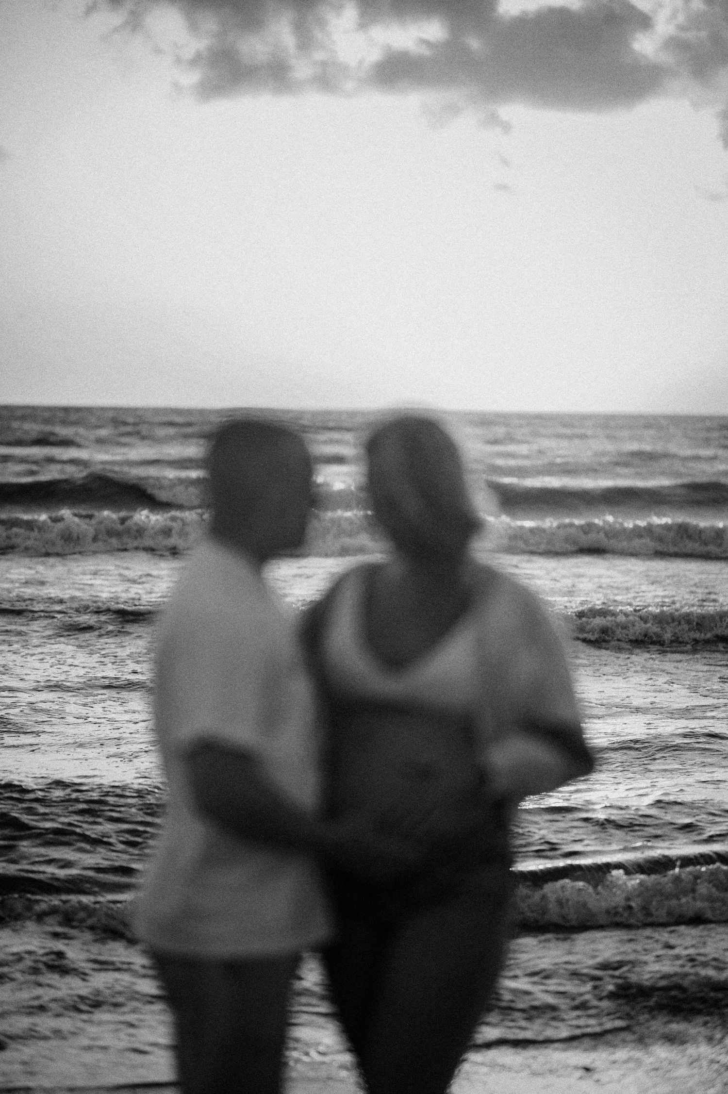 A silhouetted couple stands together watching waves crash on a beach at dusk in black and white.