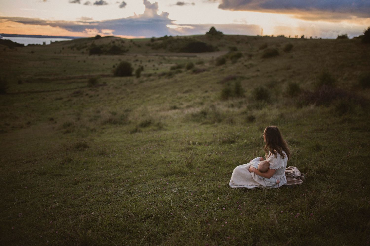Person in white dress sits alone on grassy hillside during sunset.