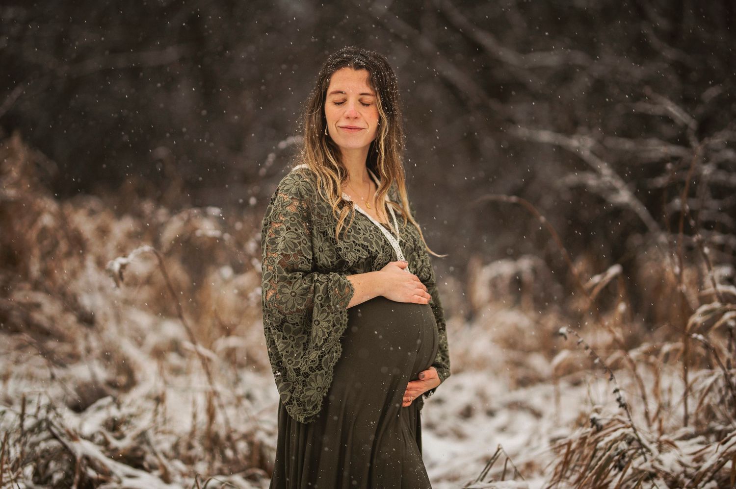 Maternity photo in winter forest showing woman in green lace dress cradling baby bump during gentle snowfall.