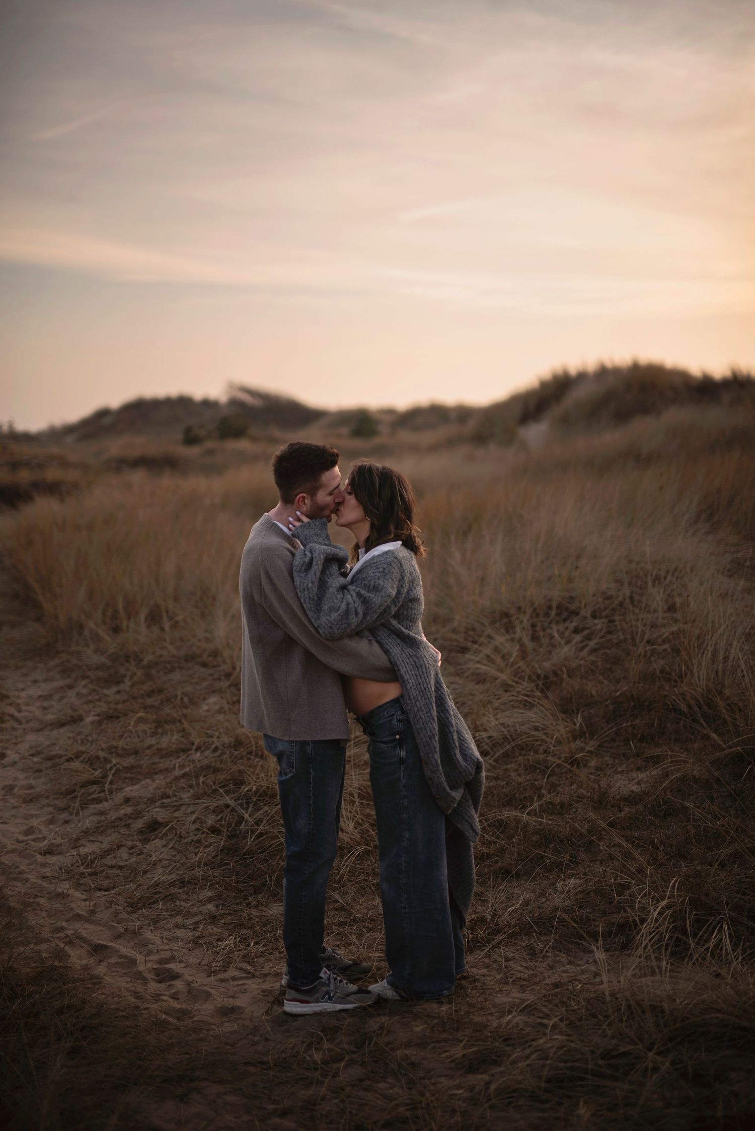 A couple shares a romantic moment at sunset among tall grasses on a hillside path.