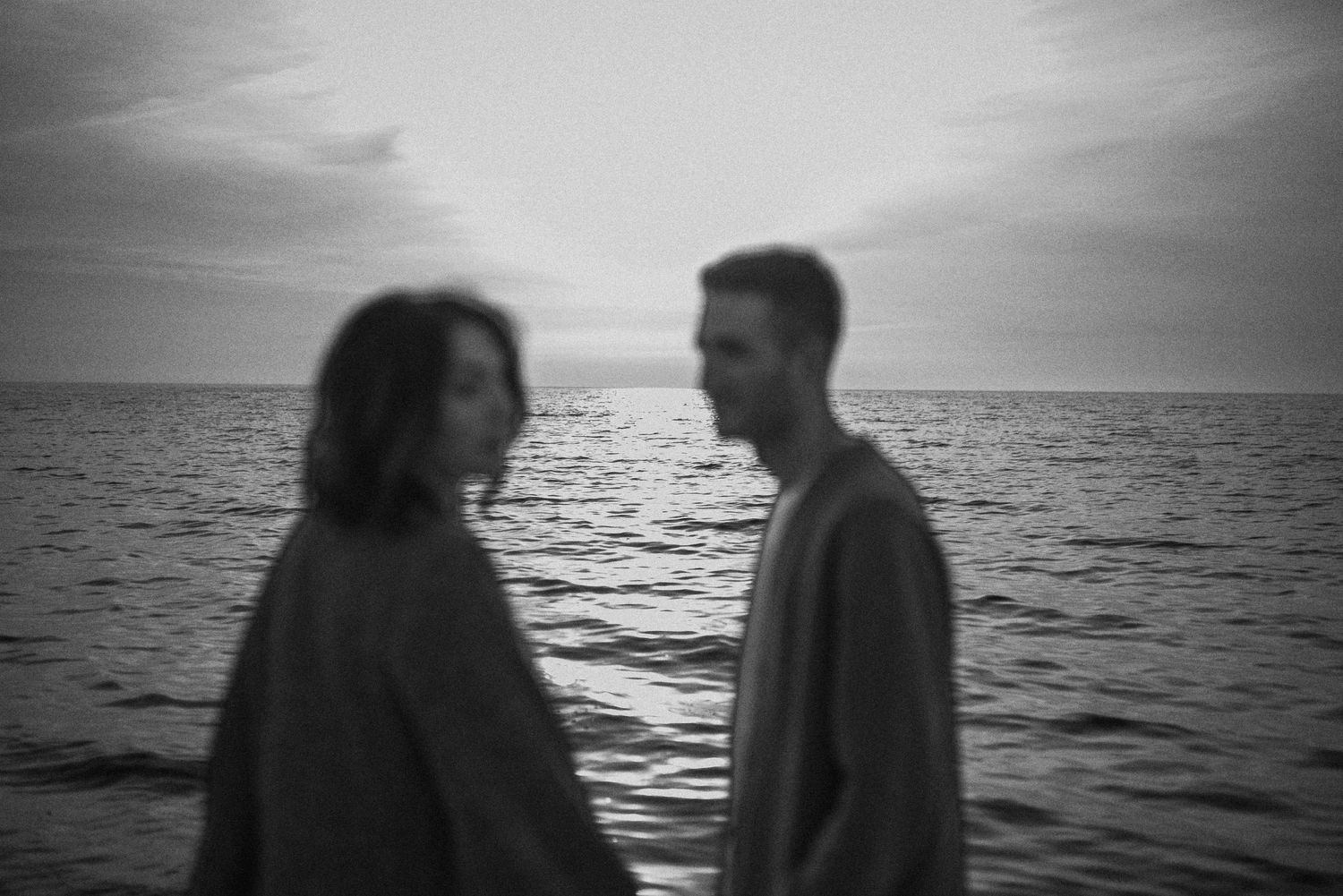 Black and white silhouette of a couple in profile standing near the ocean at sunset.