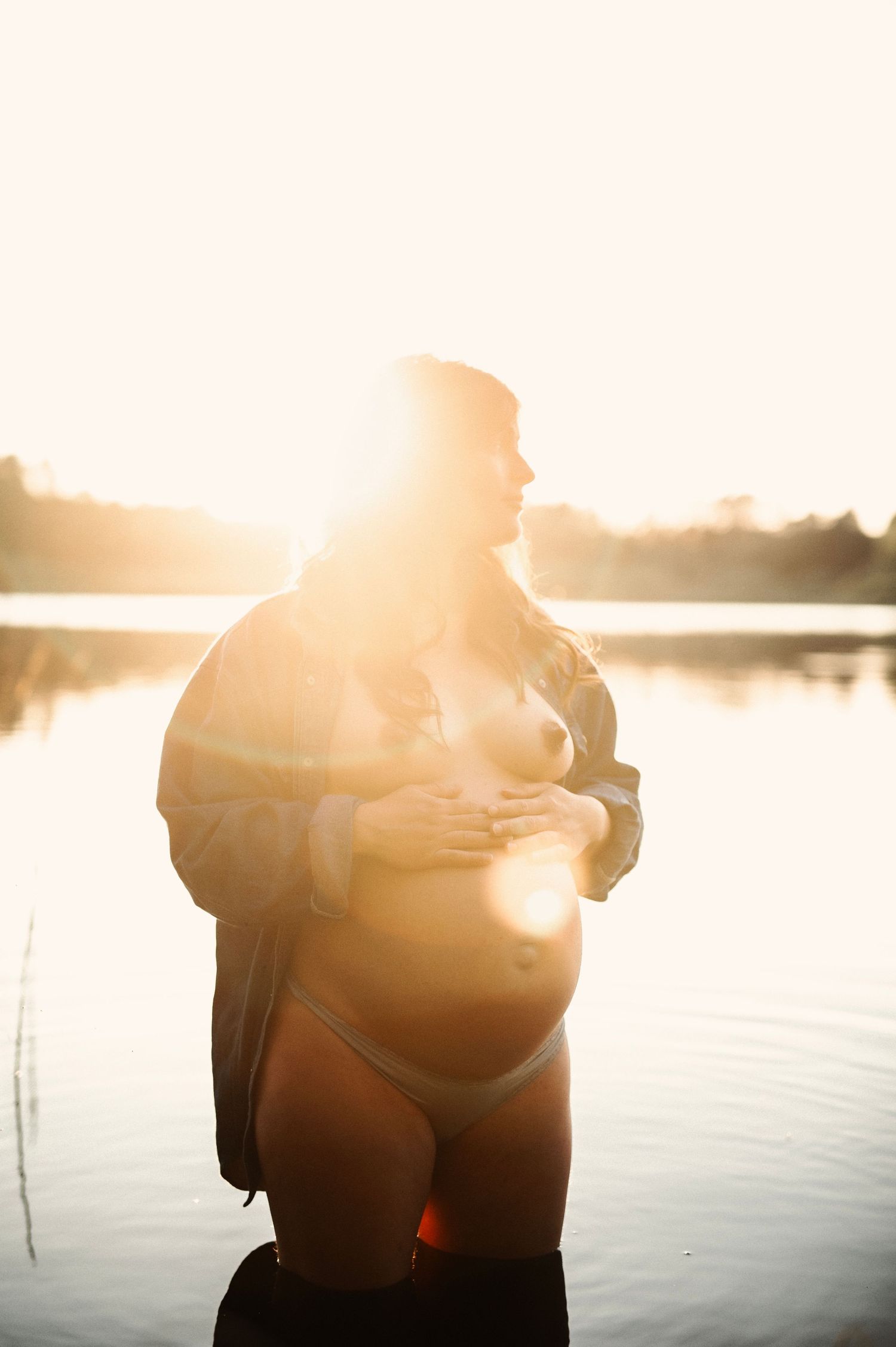 Sunlit maternity portrait by a serene lakeside during golden hour.