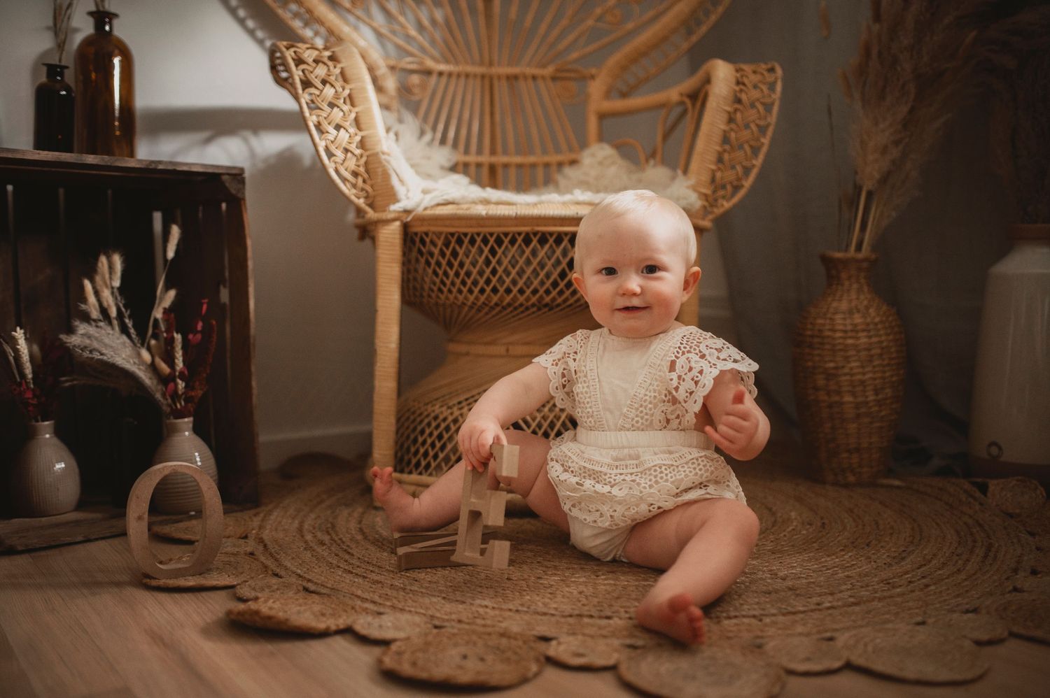 Baby in vintage lace outfit sitting on round mat with boho decor and wicker chair.