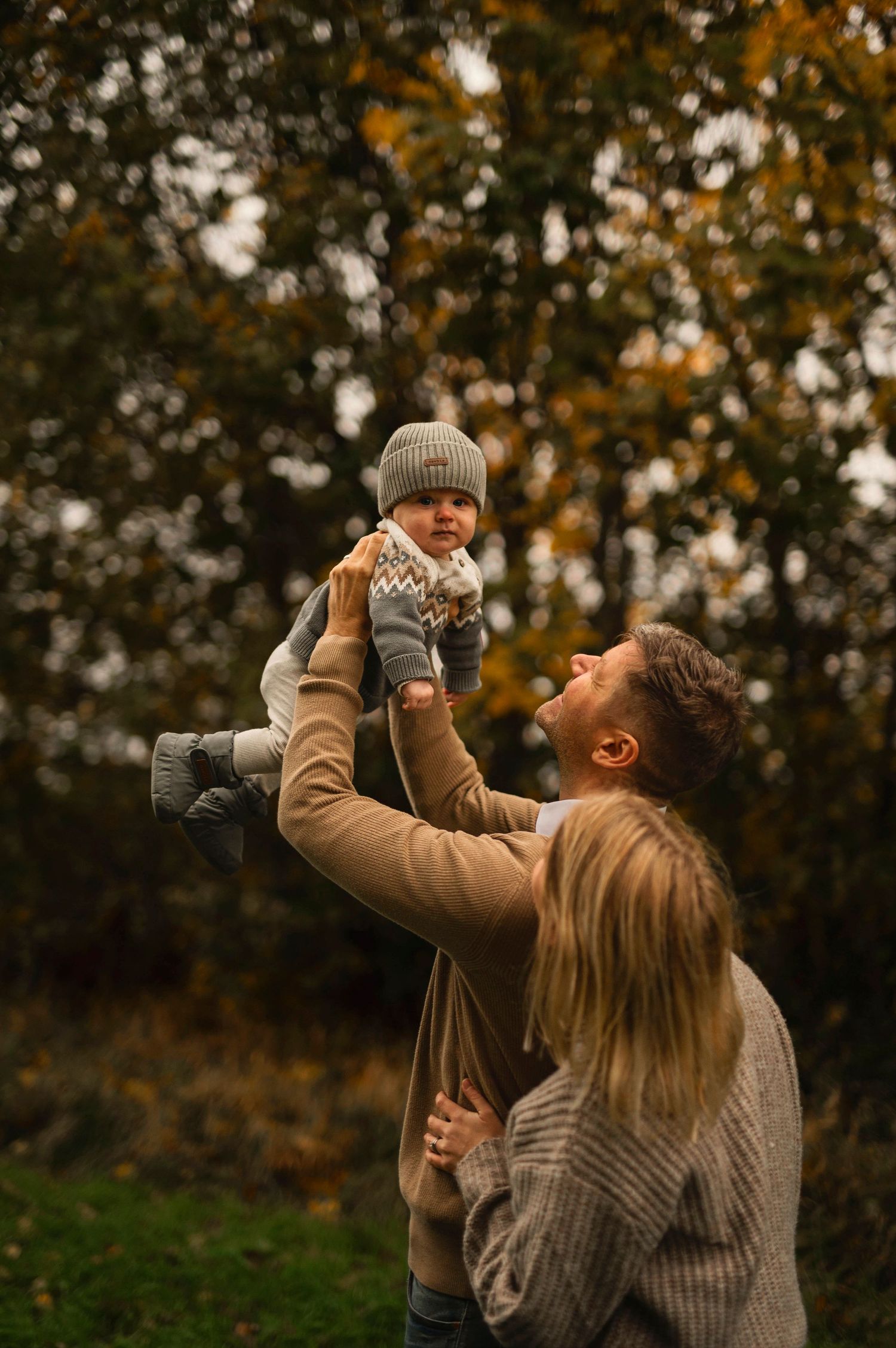 A baby in a grey hat is lifted up against fall foliage.