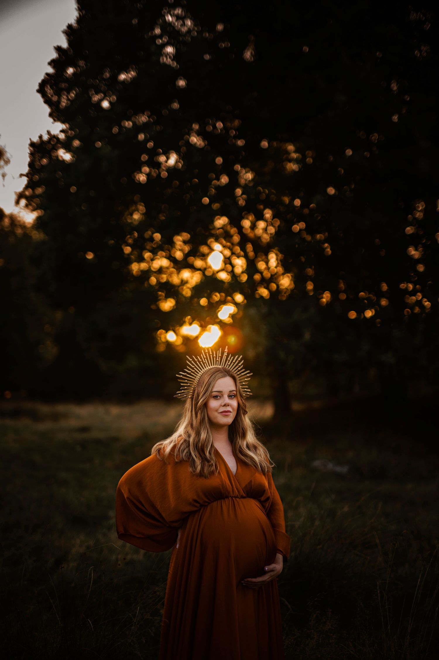 Dreamy sunset maternity photo with crown detail and flowing rust-colored dress in natural setting.