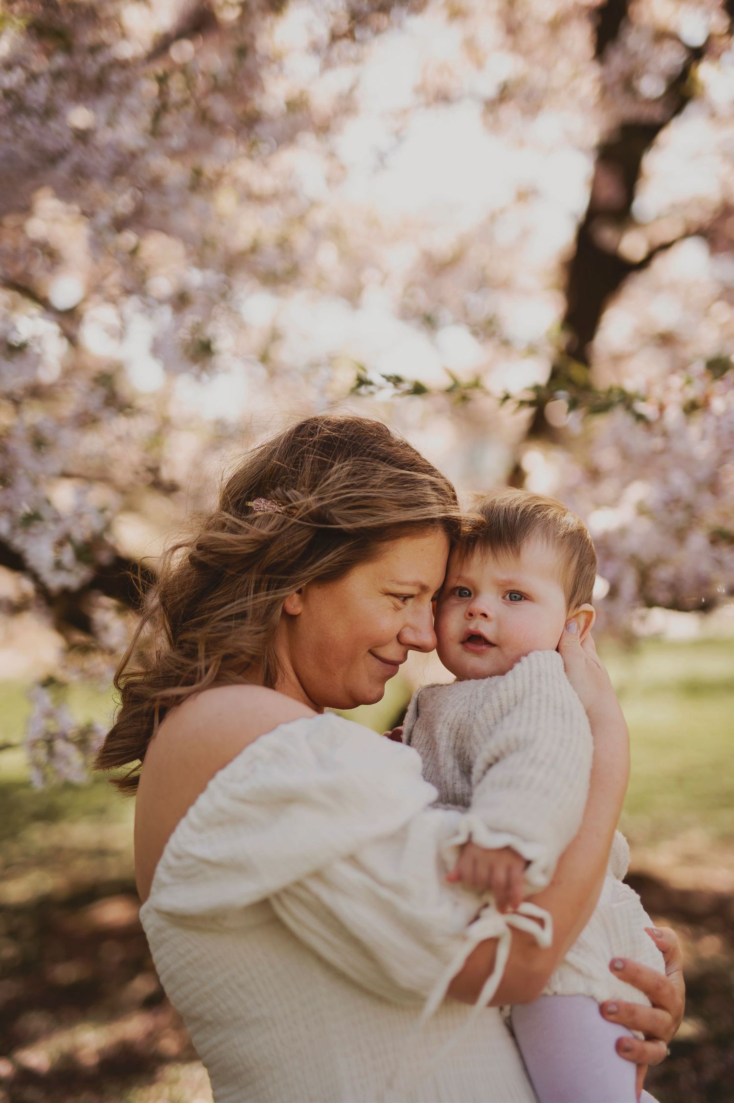 A mother in a white dress embraces her baby among cherry blossoms during a sunset photo shoot.