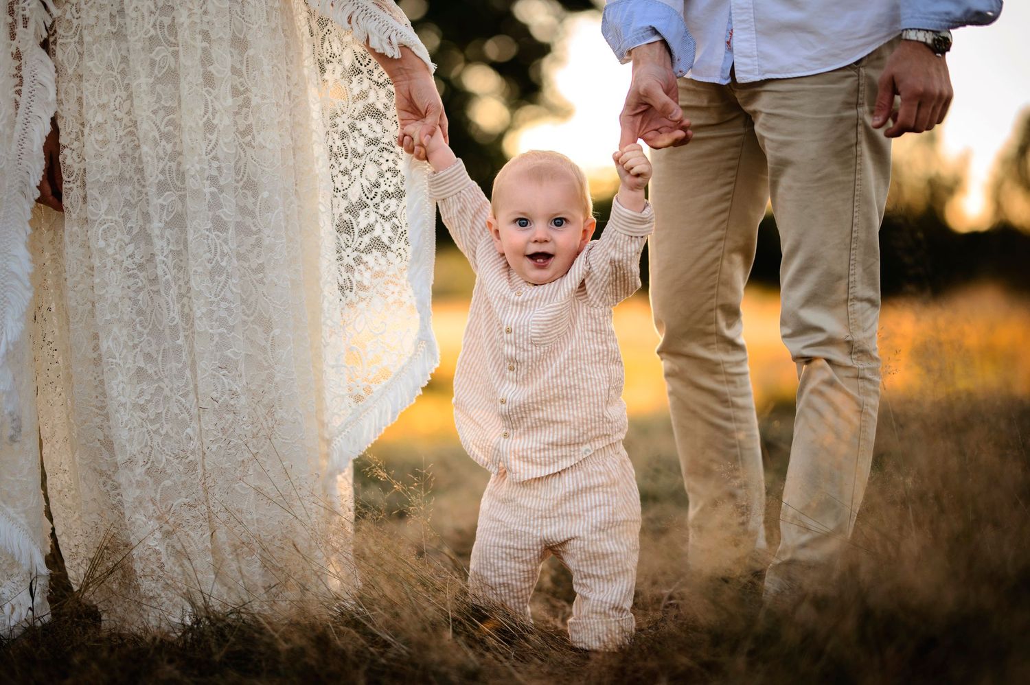 A joyful baby in white clothing learns to walk outdoors at sunset.
