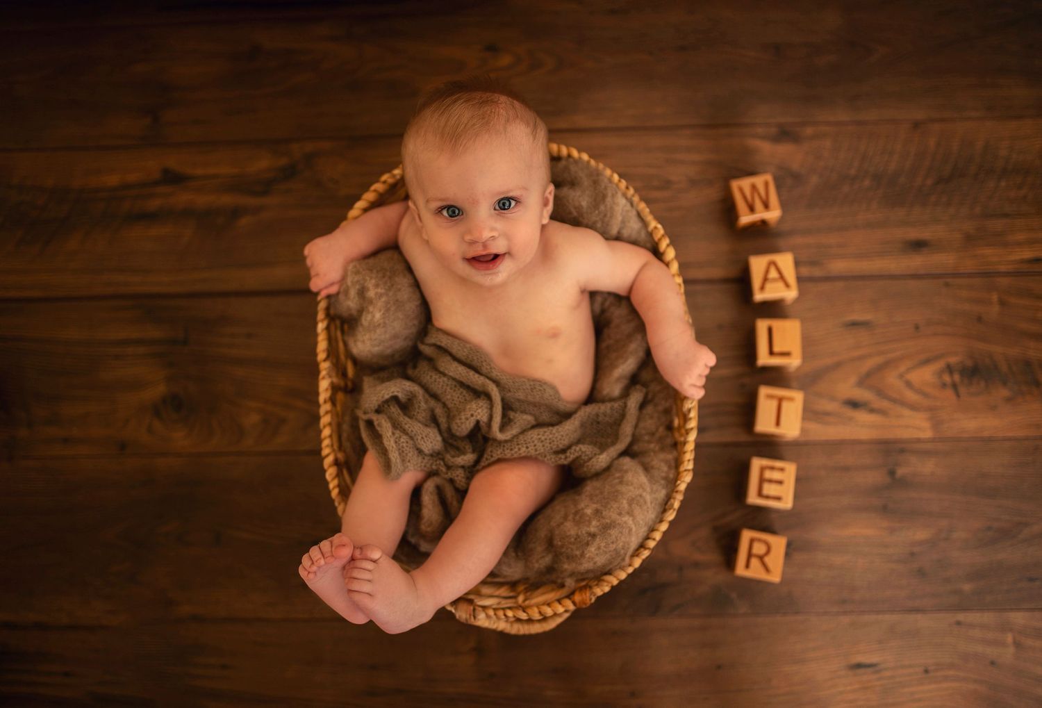 Happy baby in basket with wooden letter blocks on dark wooden floor.