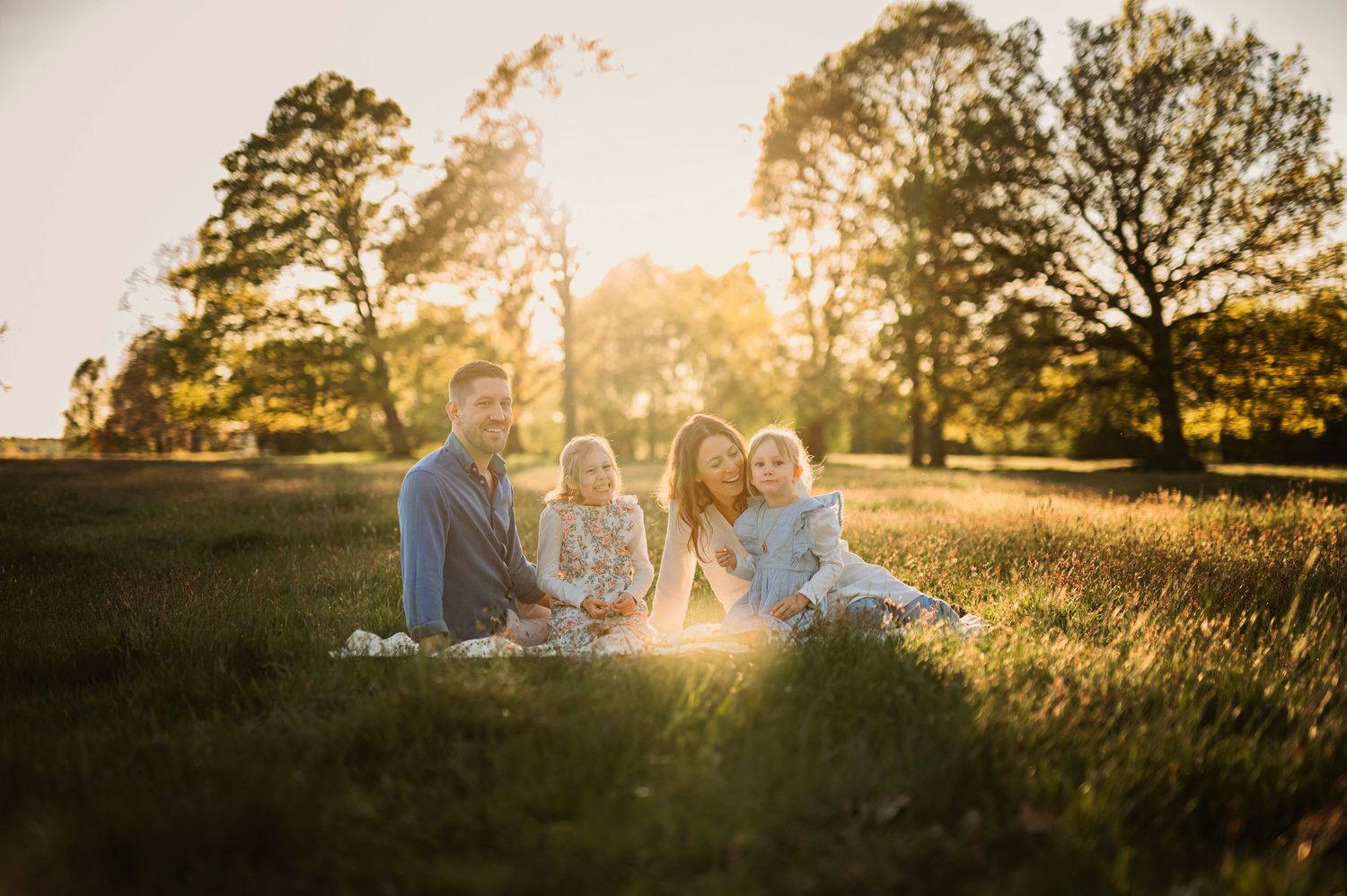 Family having a picnic in golden sunset light on grassy field with tall trees in background.