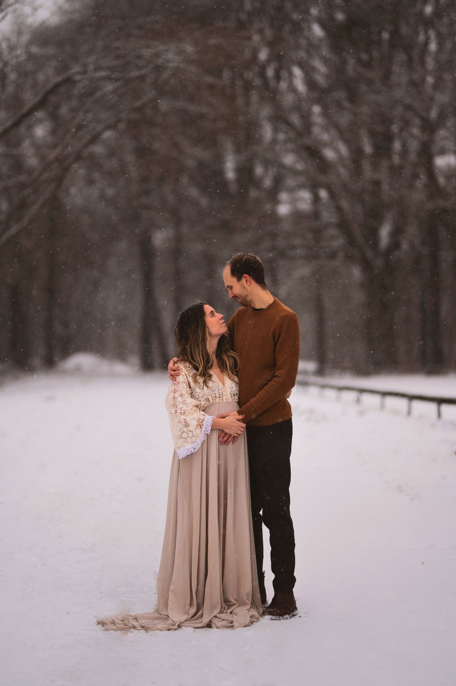 A romantic winter photoshoot features a couple in warm attire standing close together in a snowy forest setting.