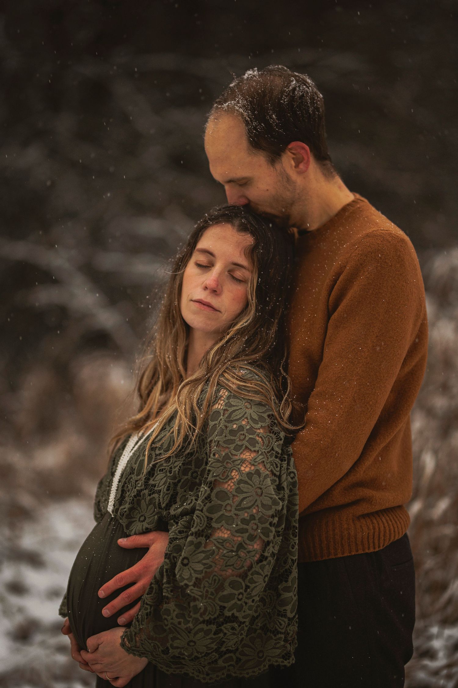 Tender moment captured of expecting couple in winter scene with rust sweater and lace dress.