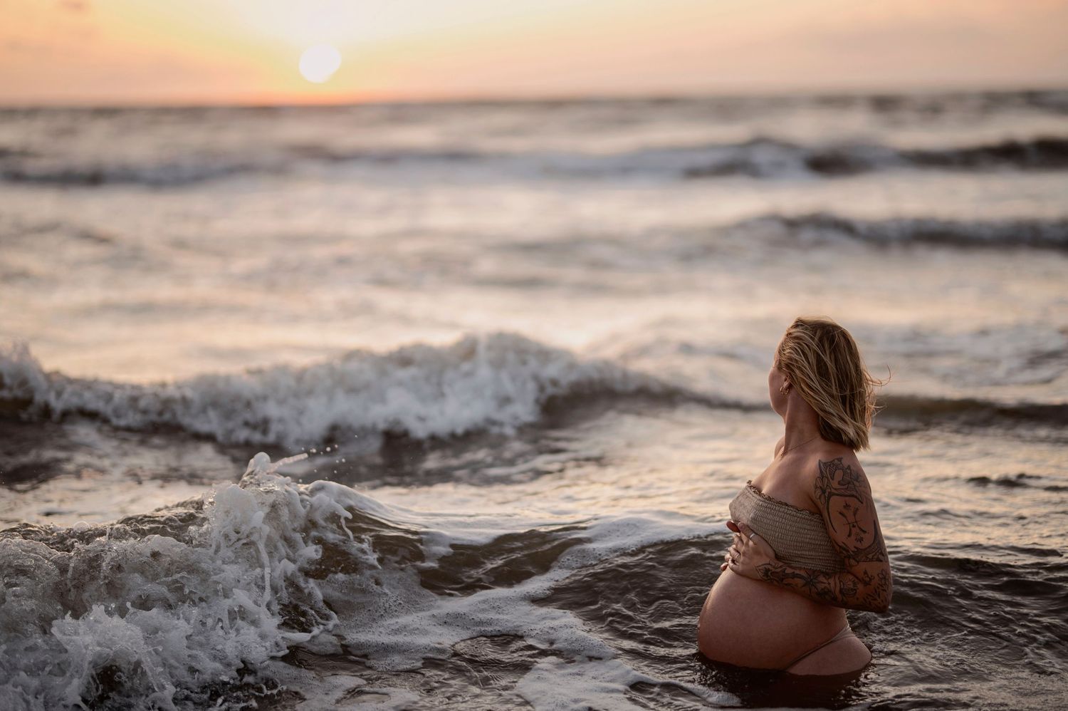 A figure sits in churning ocean waves at sunset while golden light illuminates the dramatic seascape.