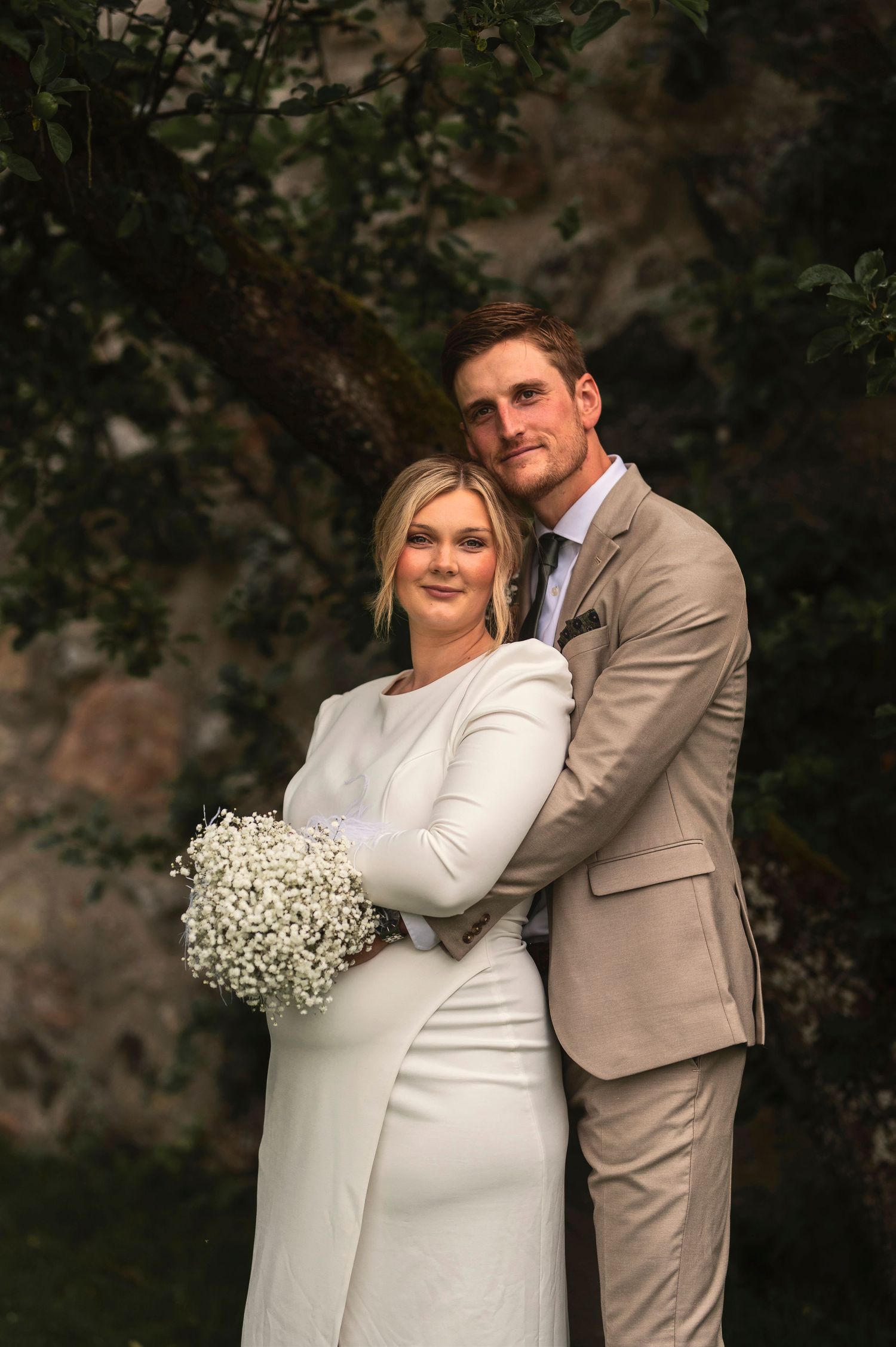Wedding couple poses against garden backdrop with white floral bouquet.