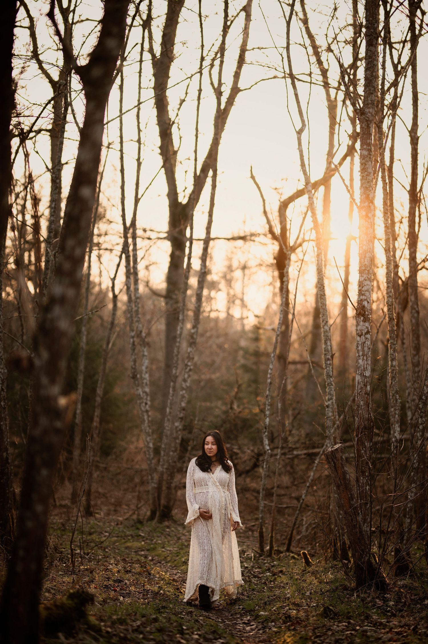 Ethereal figure in a flowing white dress stands among bare trees at sunset in a misty forest.