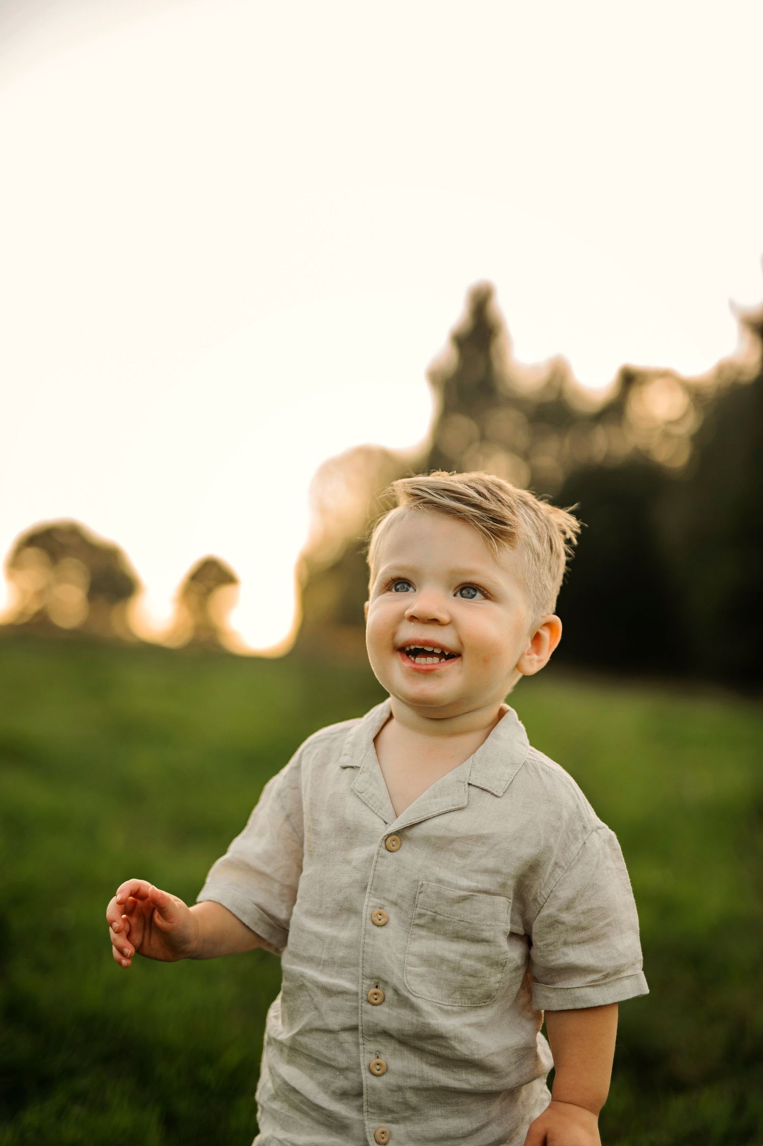 A joyful toddler in a beige button-up shirt smiles during golden hour outdoor photography.