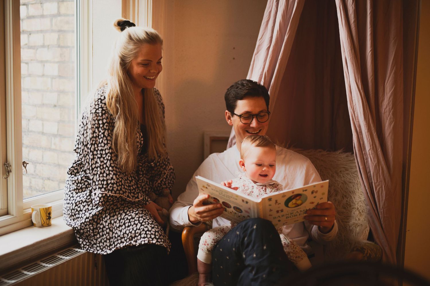 Young family with baby enjoying storytime together by sunny window at home.