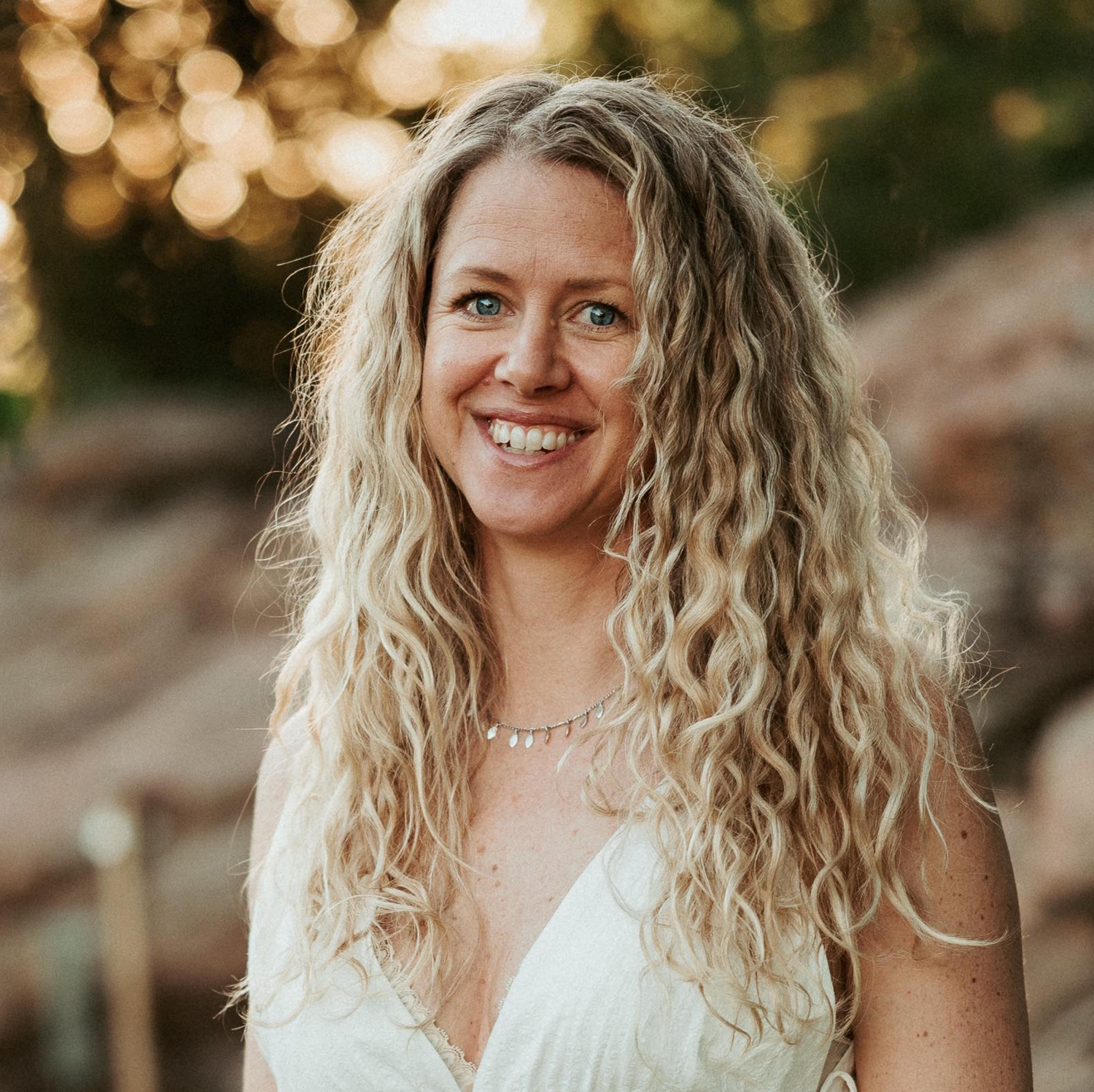 Smiling blonde woman with curly hair wearing white dress outdoors at golden hour.