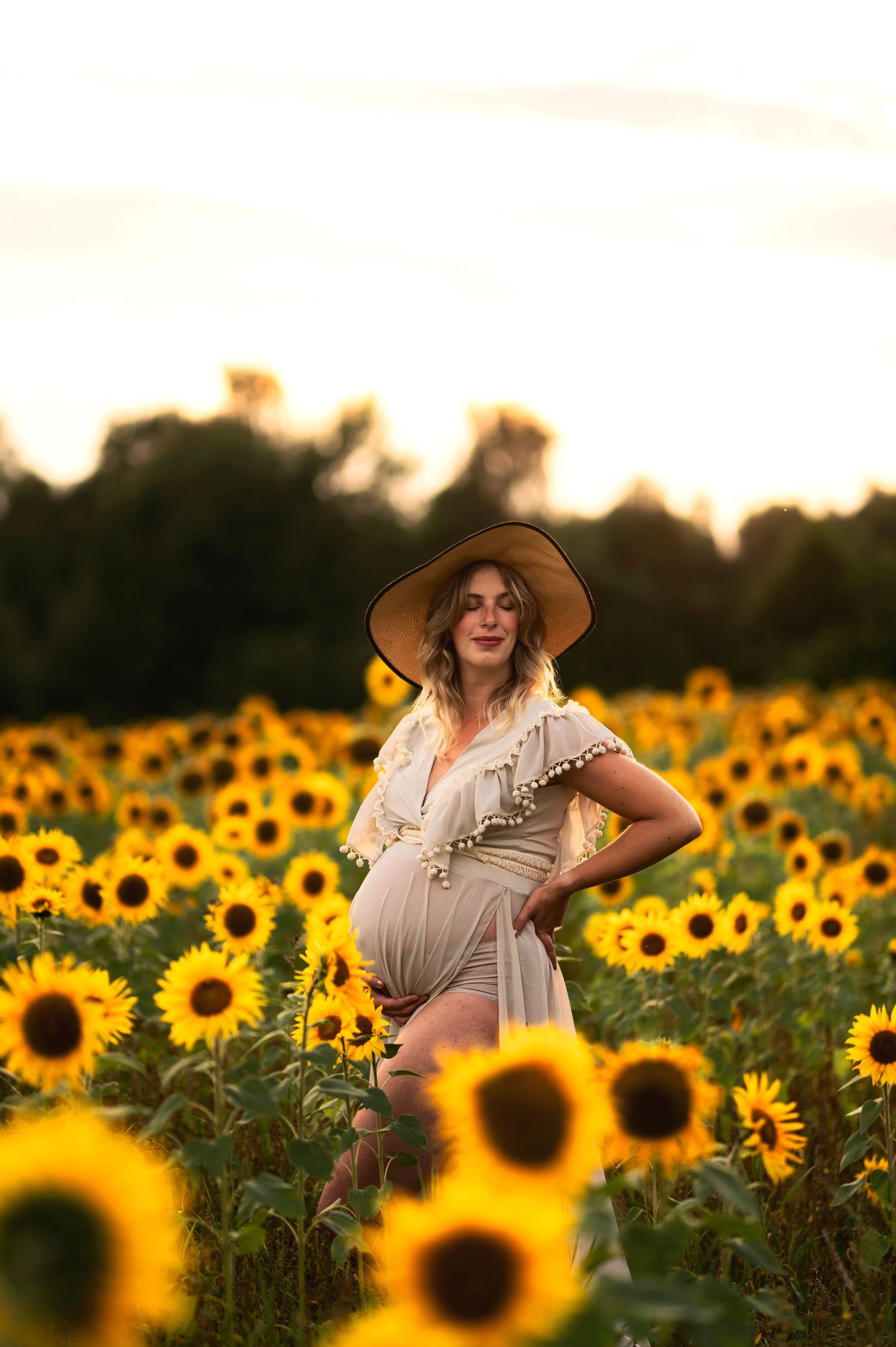 Summer maternity portrait in beige dress and hat surrounded by bright sunflowers.