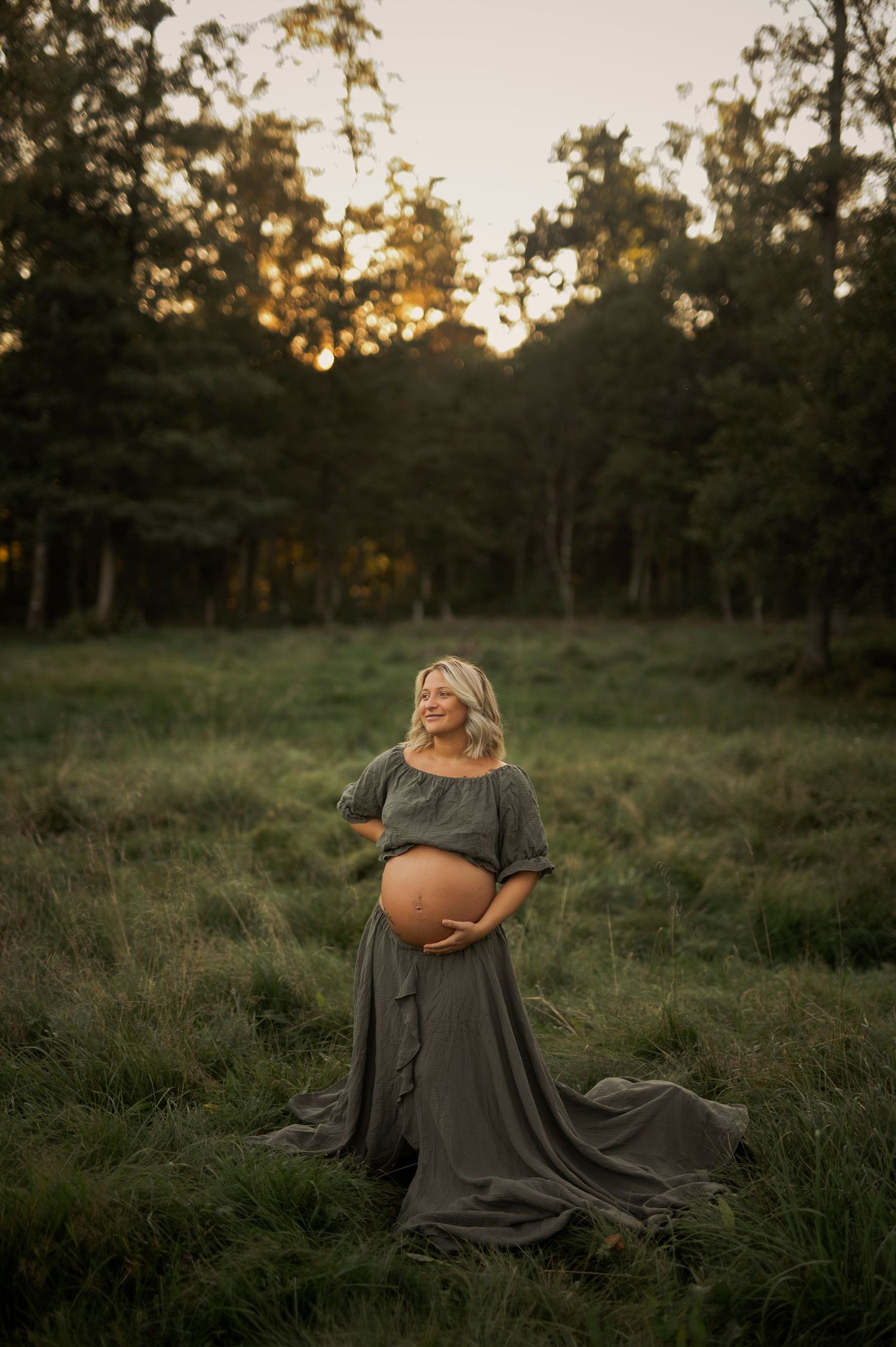 Expectant mother in flowing gray dress poses gracefully in a sunlit meadow at golden hour.