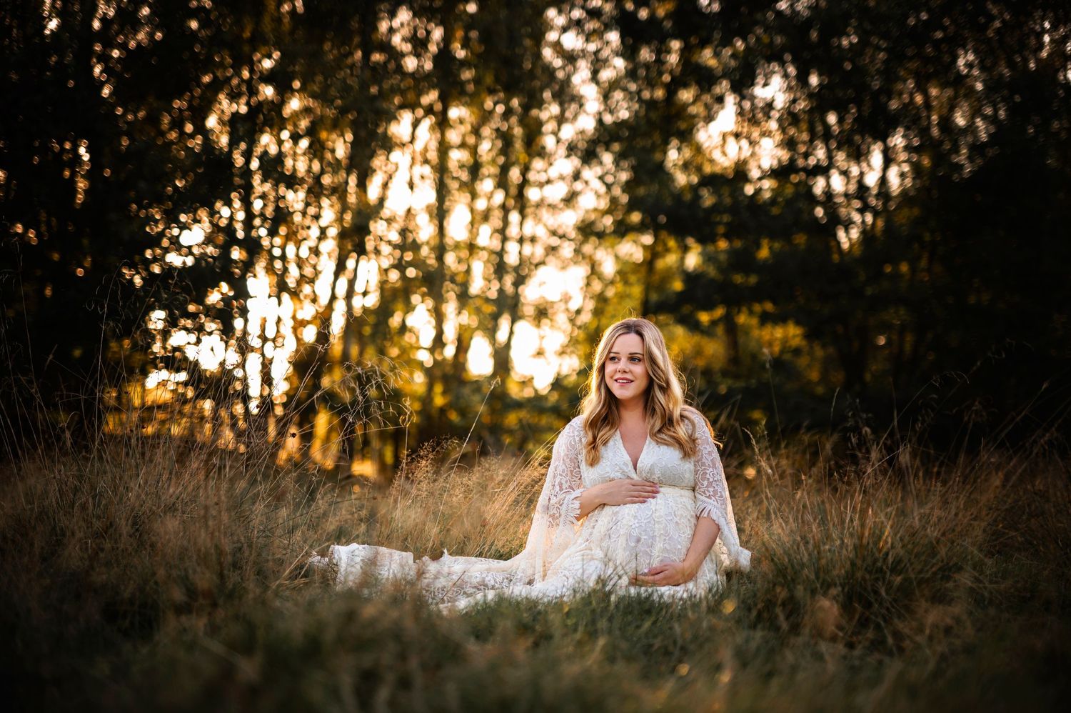 Glowing maternity portrait in white dress amid sunlit field at dusk.