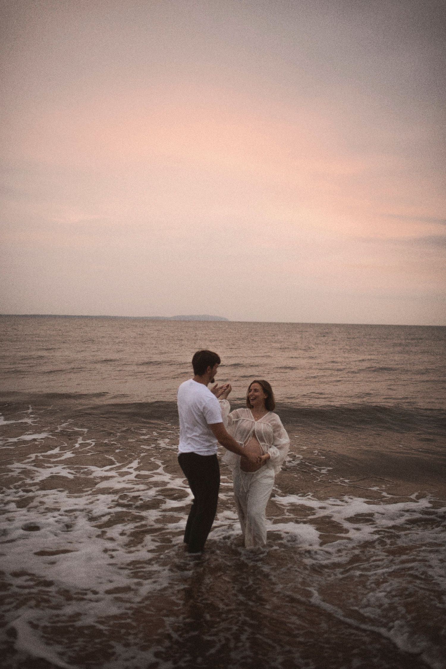 A couple wades in the ocean waves at dusk during a romantic beach photoshoot.