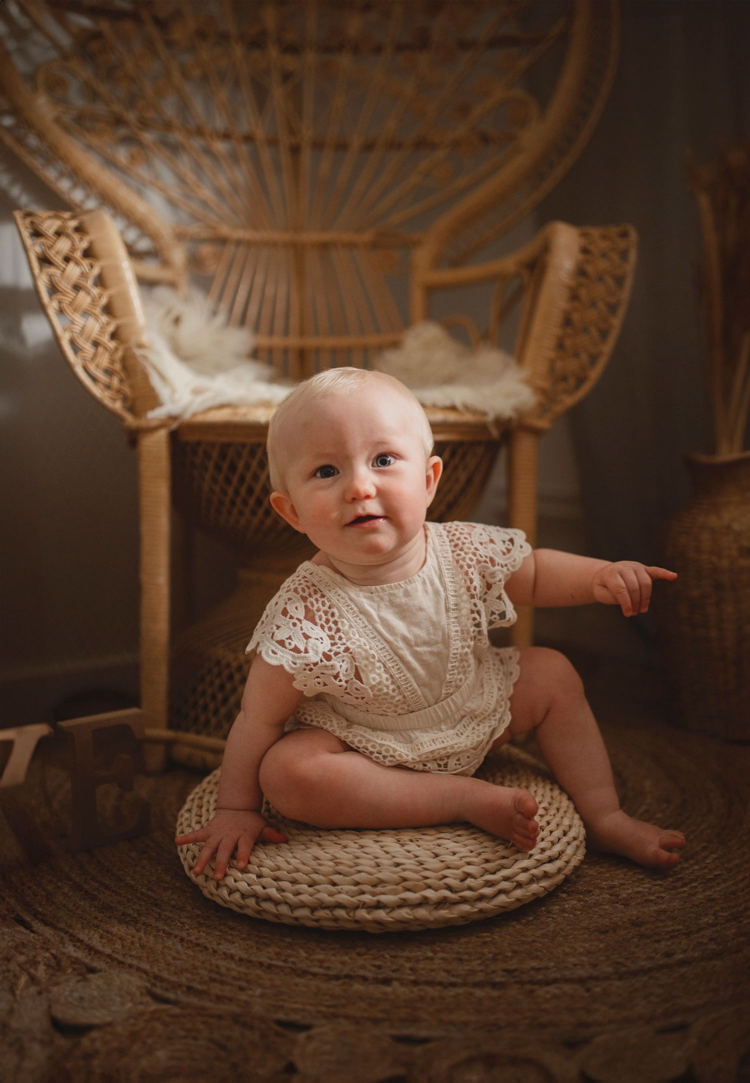 Baby in white lace dress sitting on woven mat with peacock chair in background.