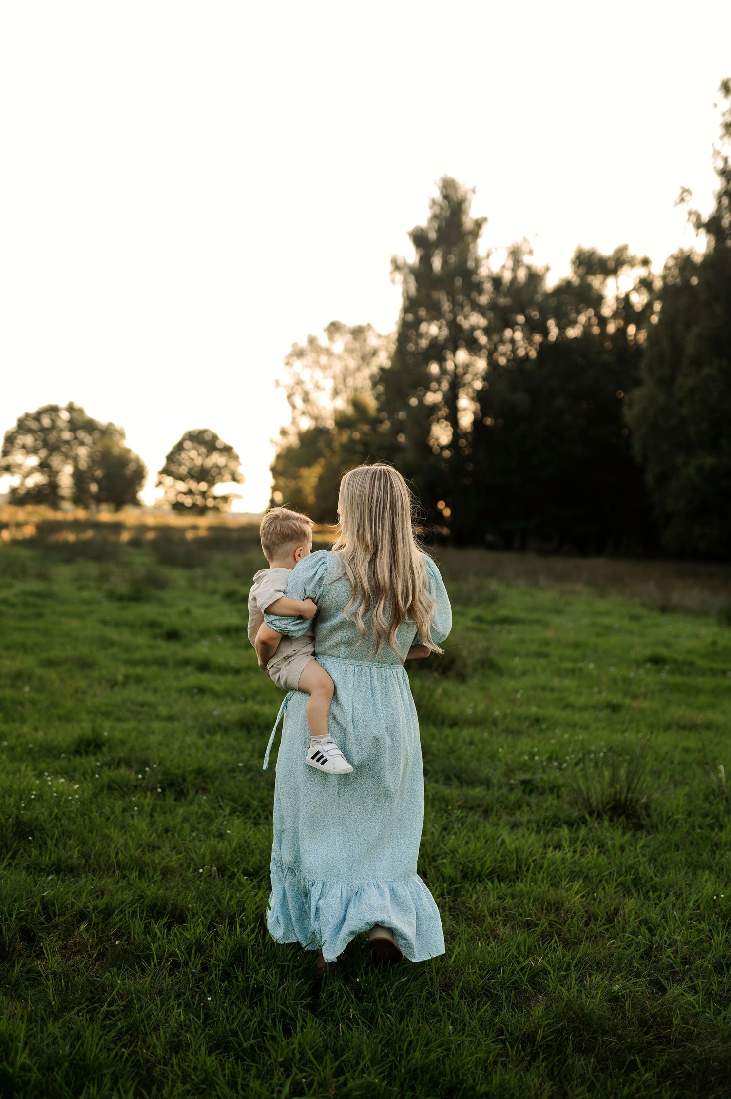 A person in a flowing blue dress holds a small child while standing in a meadow at dusk.