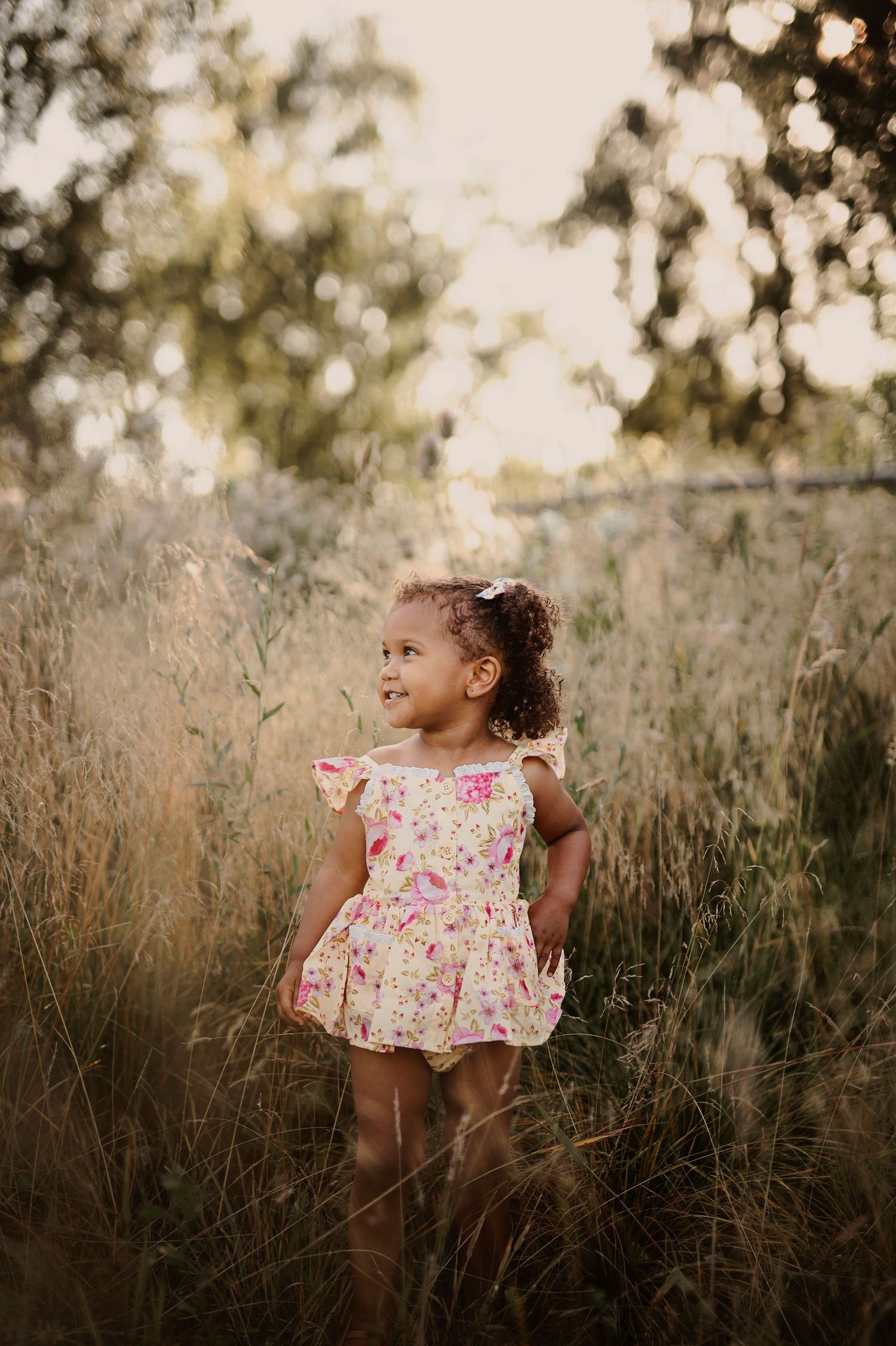 A child in a floral dress stands among tall grass during sunset.