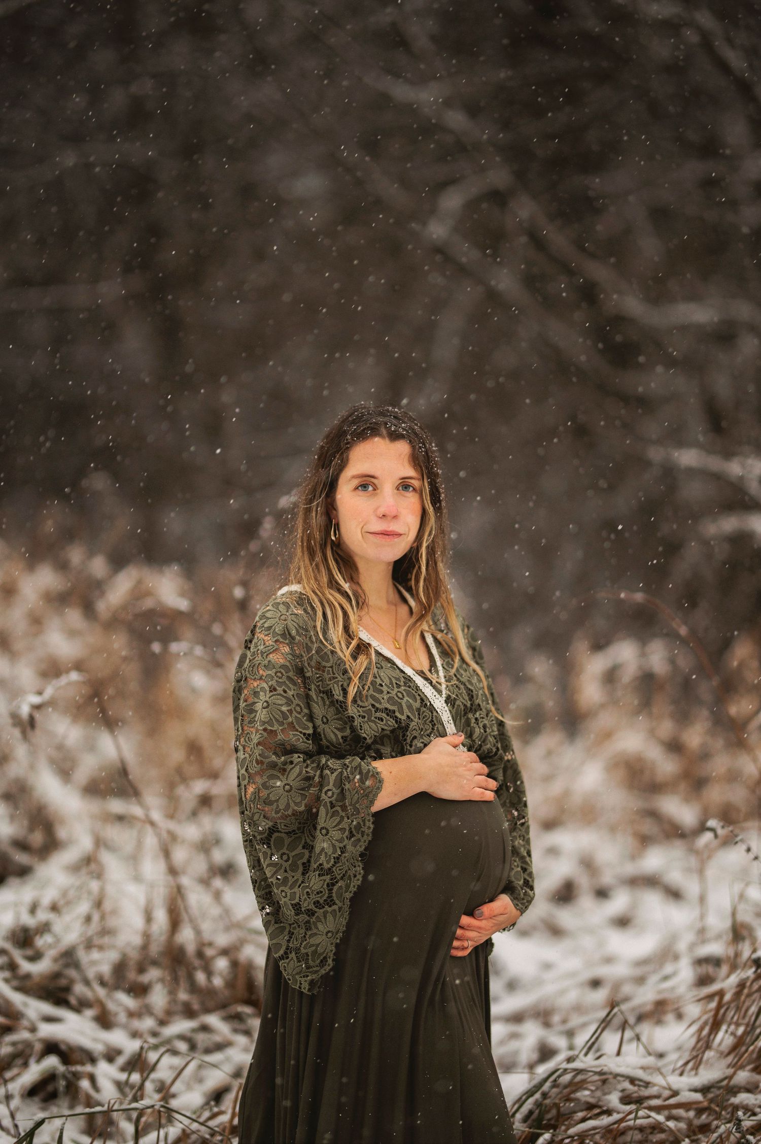 A winter maternity portrait shows an expectant mother in dark dress standing amid falling snow.