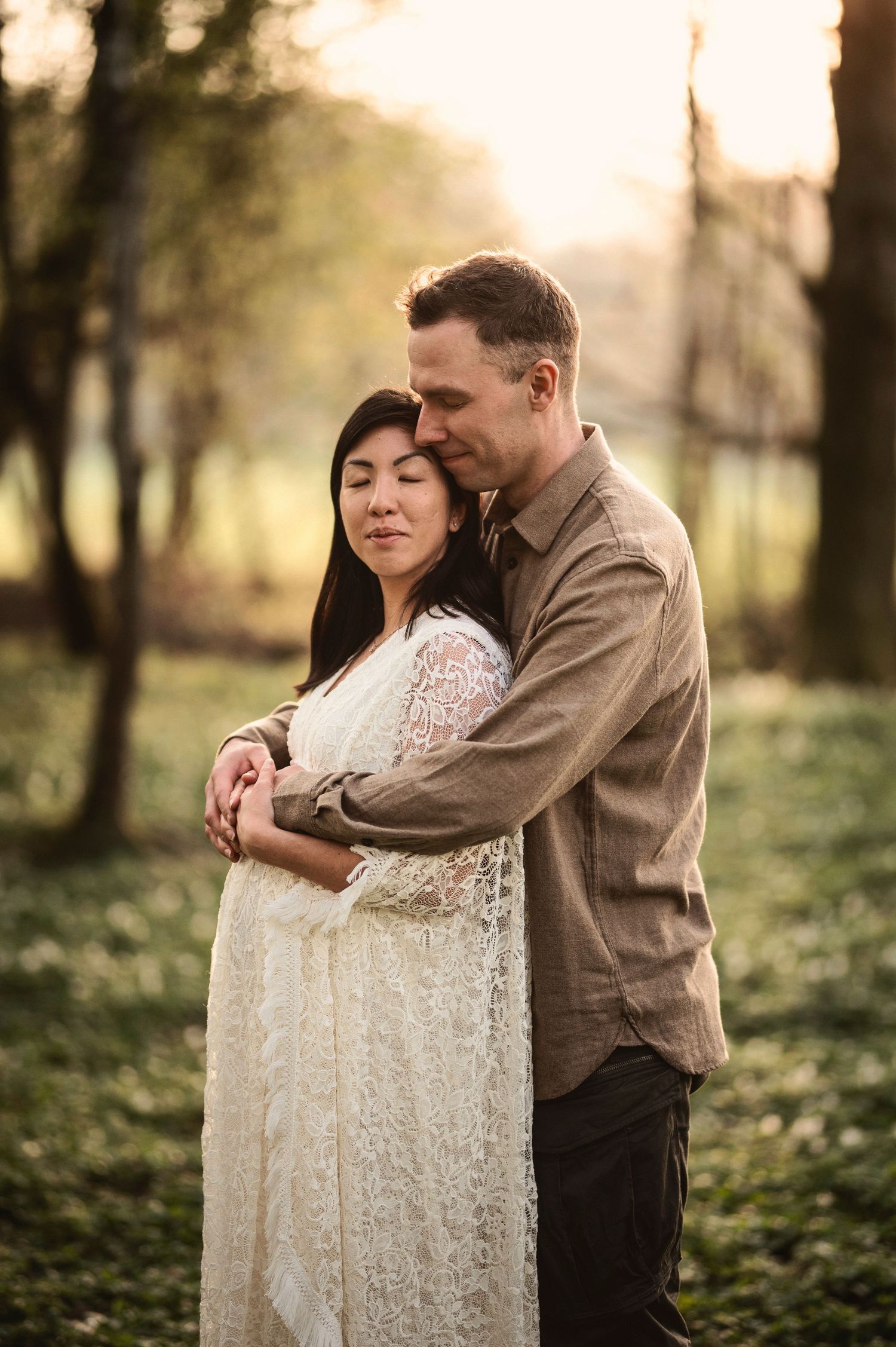 Romantic couple embraces in a dreamy forest setting at sunset with soft golden light filtering through the trees.