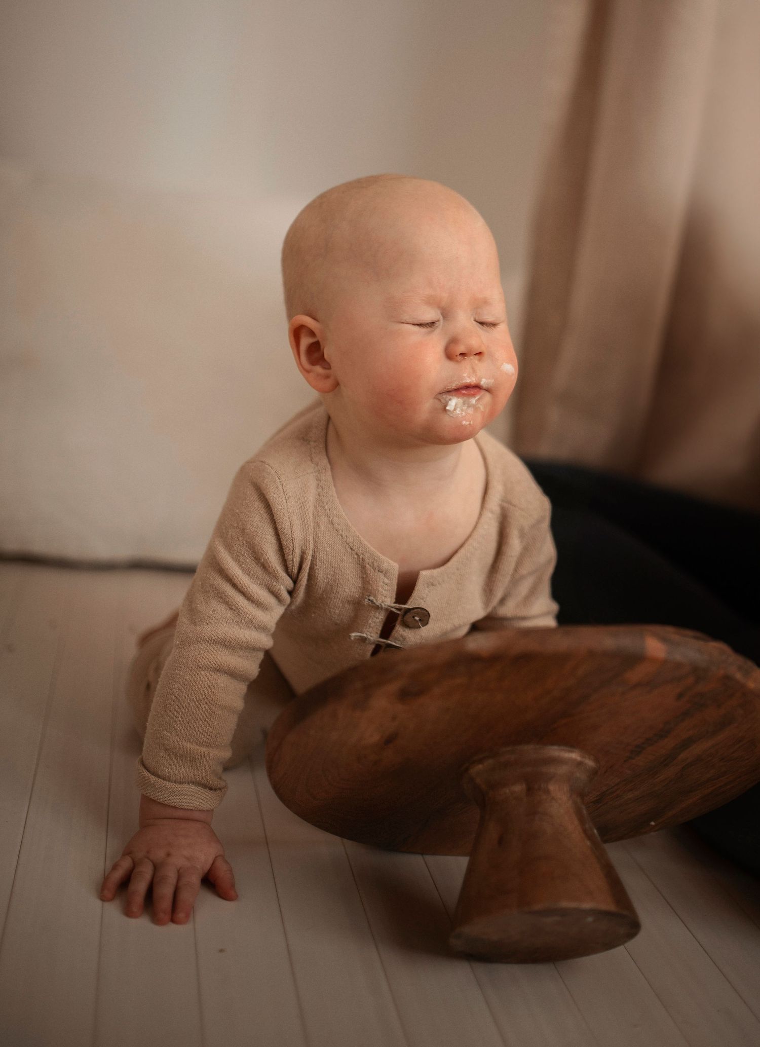 Baby in beige sweater explores wooden mushroom stool on white floor in warm lighting.
