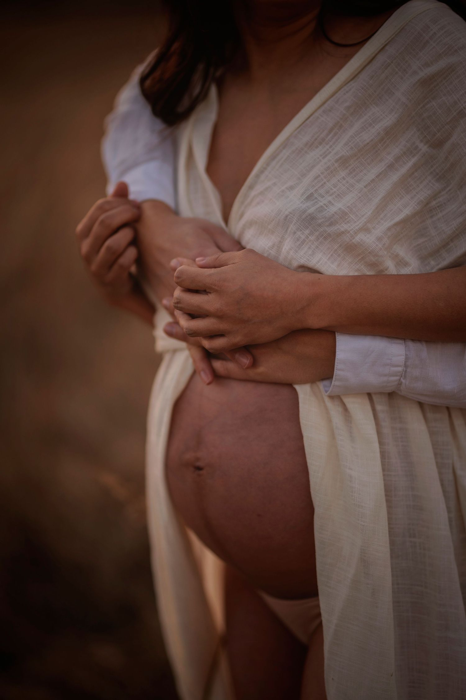 A pregnant woman in a white flowing dress holds her belly in warm natural lighting during a maternity photoshoot.