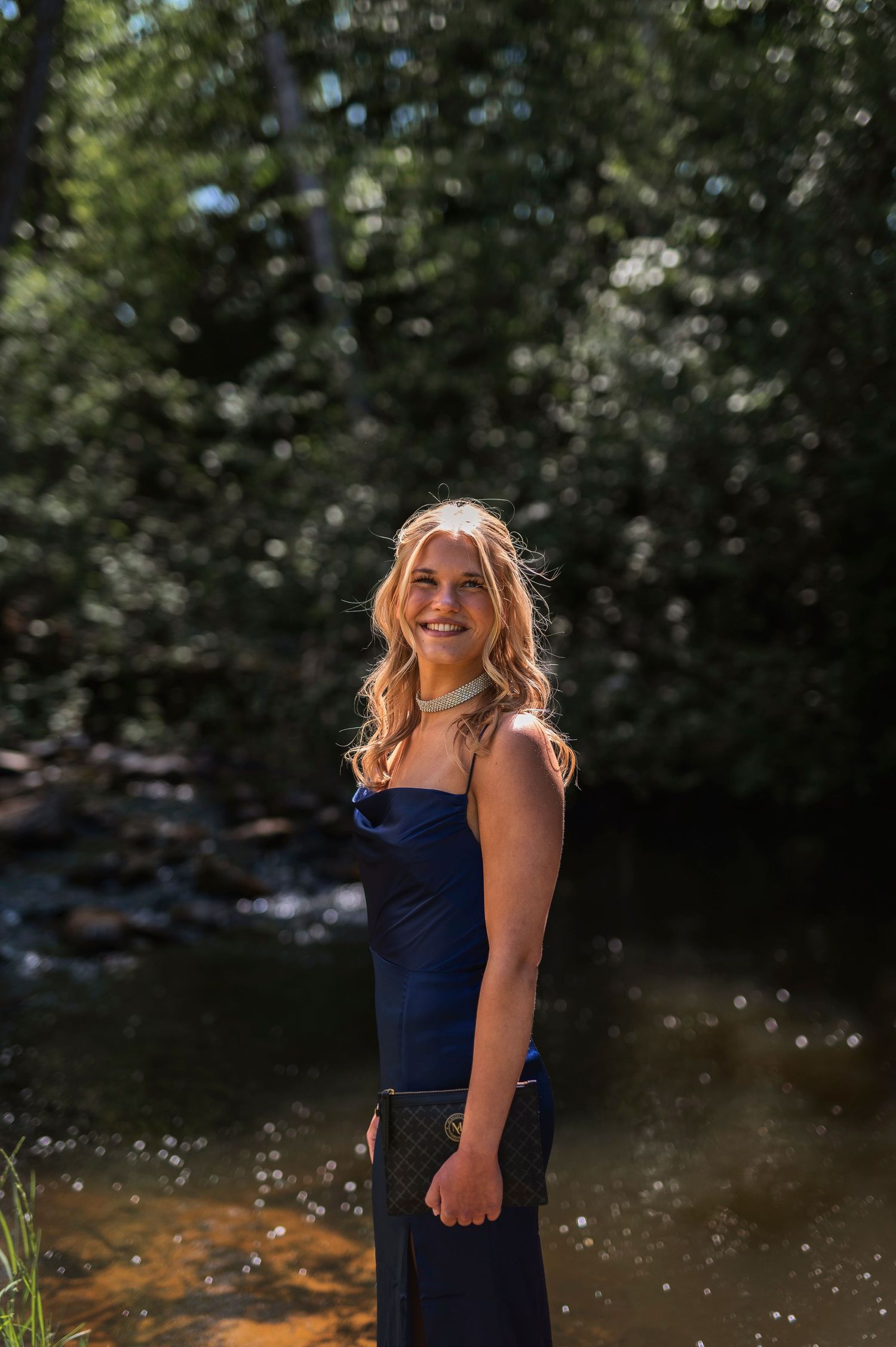 A smiling blonde woman in a navy strapless dress stands by a sunlit forest creek with dappled light.