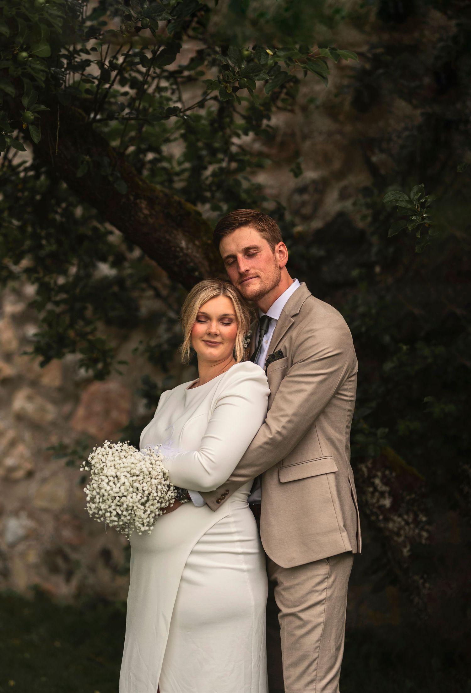 A romantic wedding portrait shows a couple embracing outdoors with the bride holding a white baby's breath bouquet.