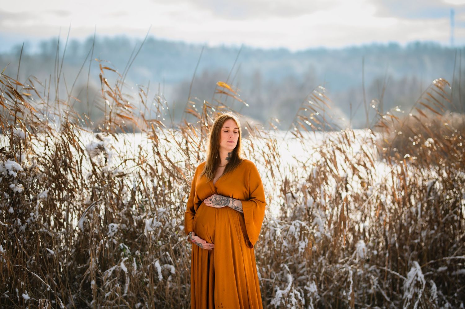 Maternity photo featuring a woman in mustard dress amid tall autumn reeds at sunset.