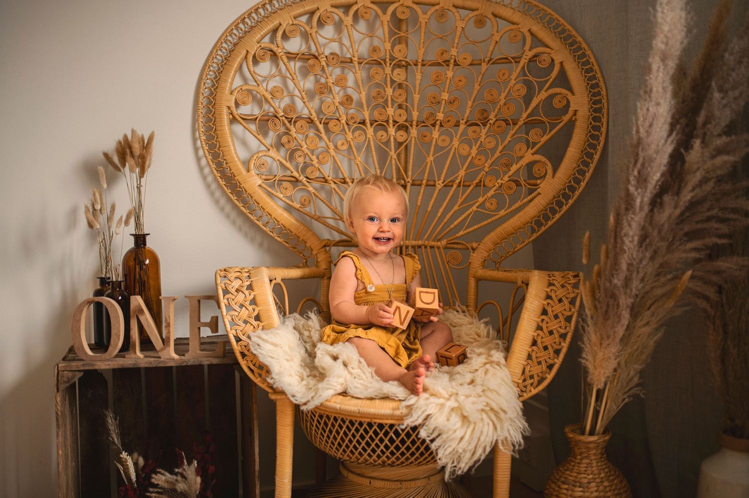 Baby sits in ornate wicker peacock chair with fluffy sheepskin and wooden ONE sign.
