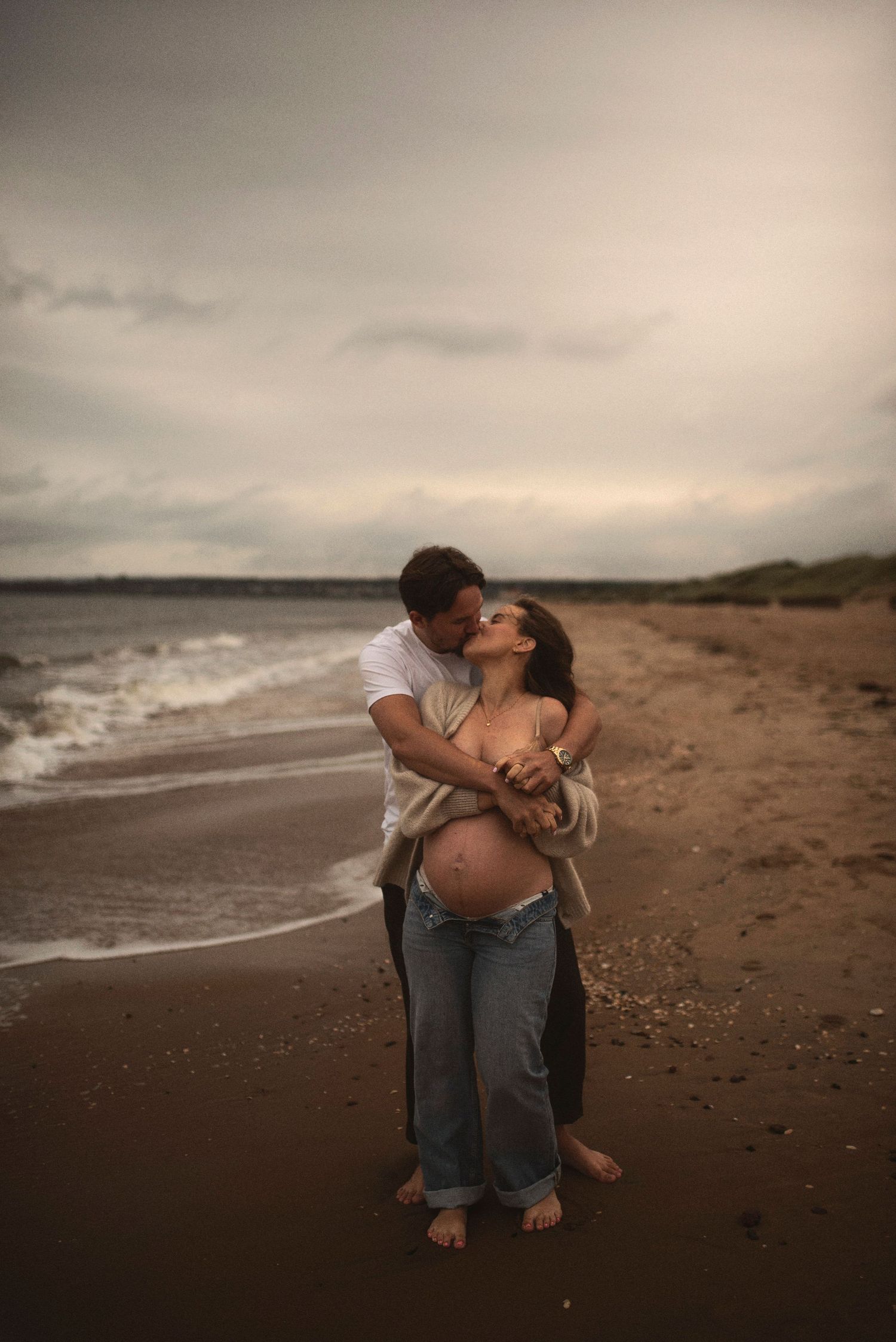 Romantic couple embraces on a moody beach at sunset with waves rolling in.