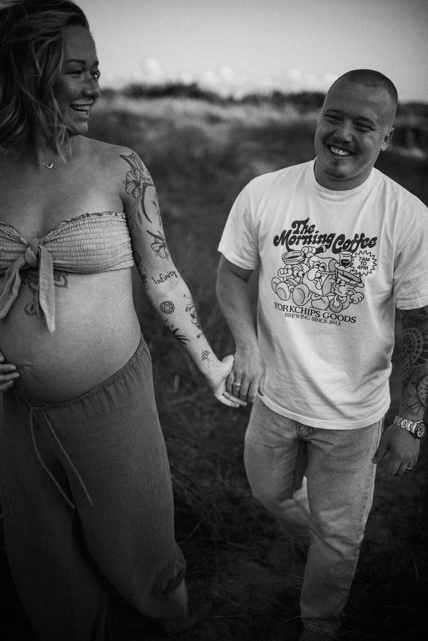A black and white photo shows a smiling couple outdoors, with a pregnant woman in a casual dress and arm tattoos.