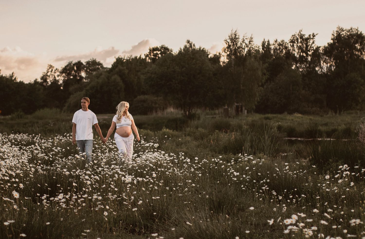 Two people walk hand in hand through a dreamy wildflower meadow at sunset with trees in the background.