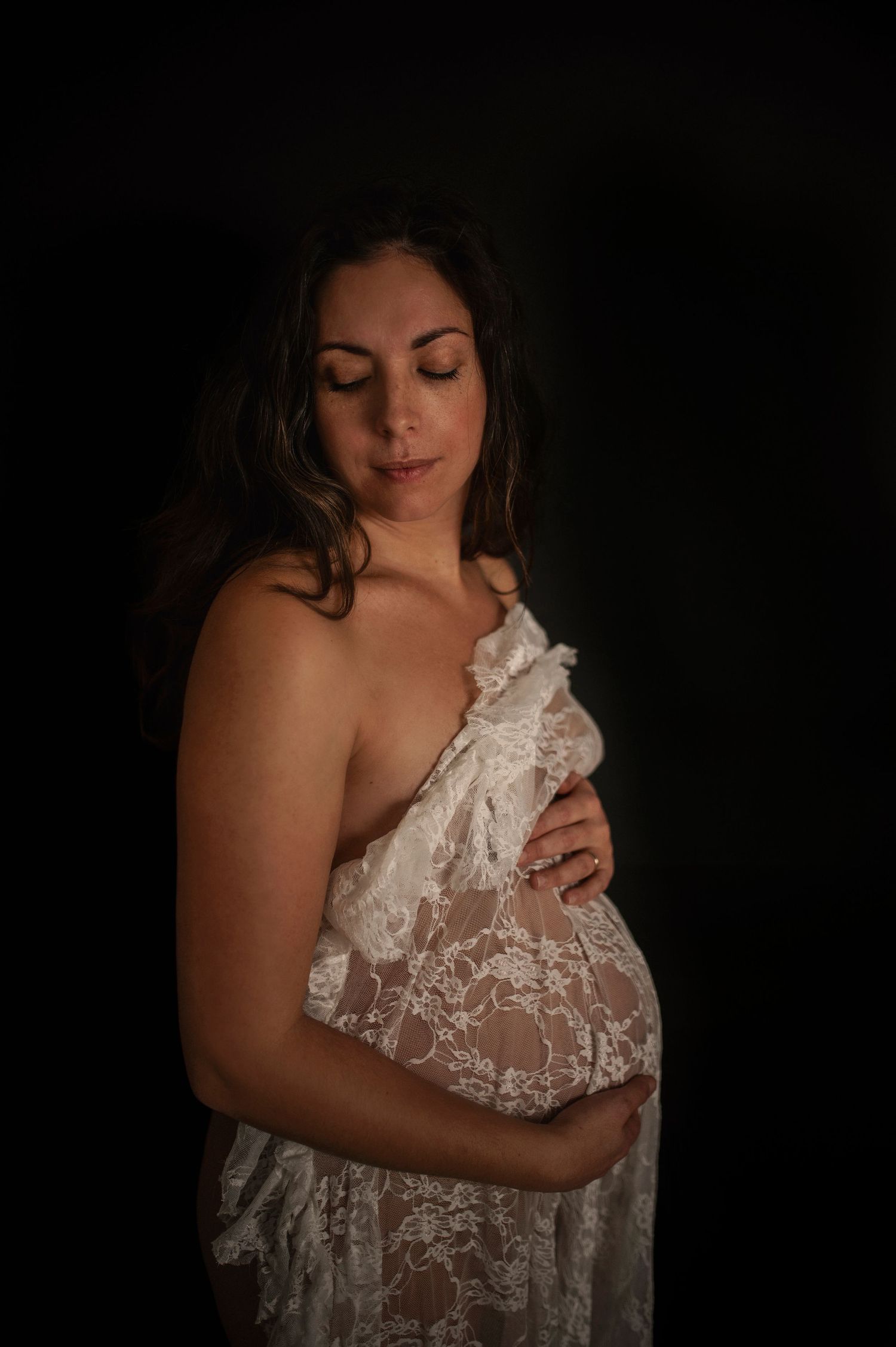 A low-key maternity portrait in dark setting with subject in white lace fabric creating an intimate, dreamy atmosphere.