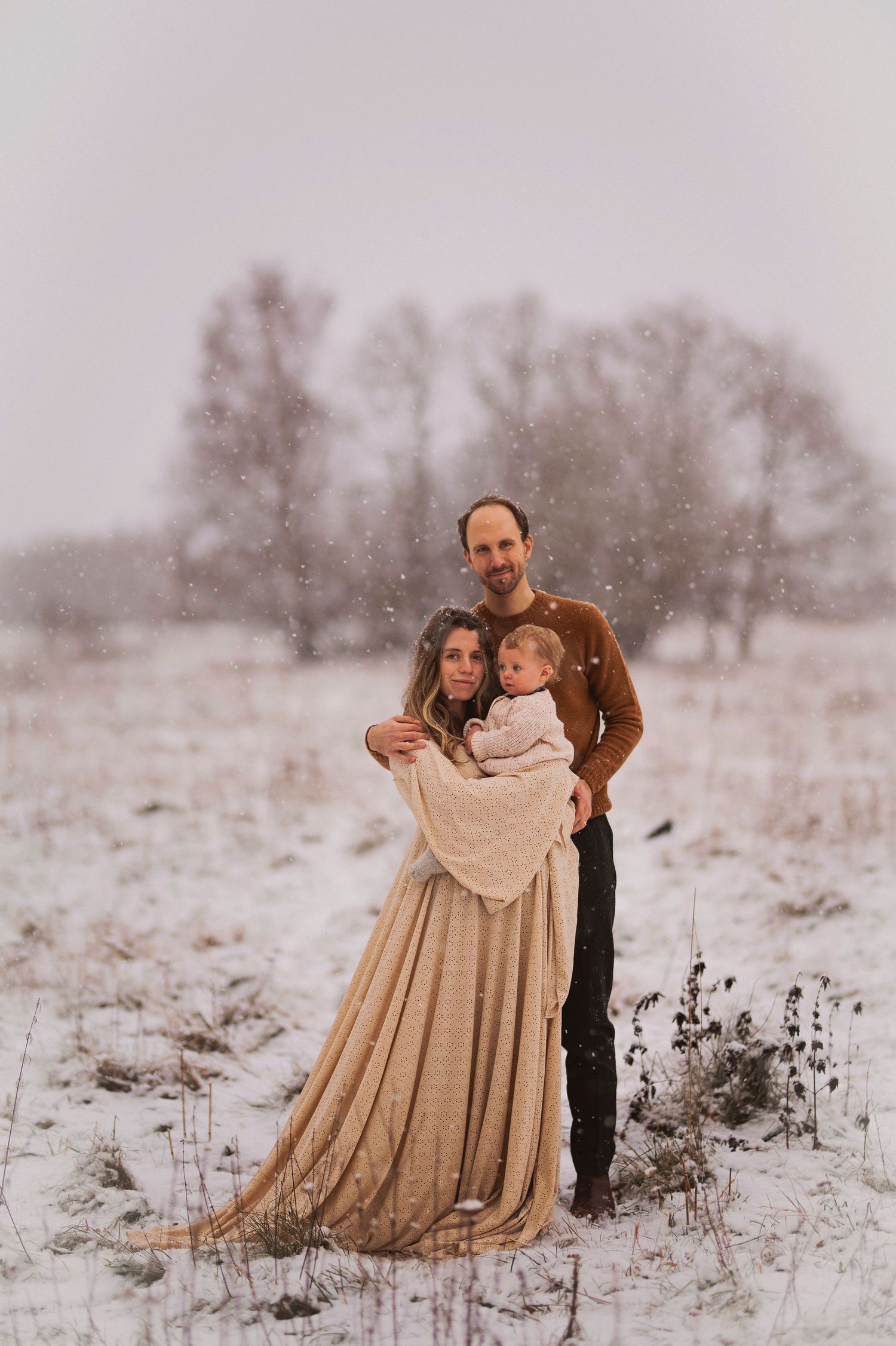 Winter family portrait in a snowy field with bare trees in the background.