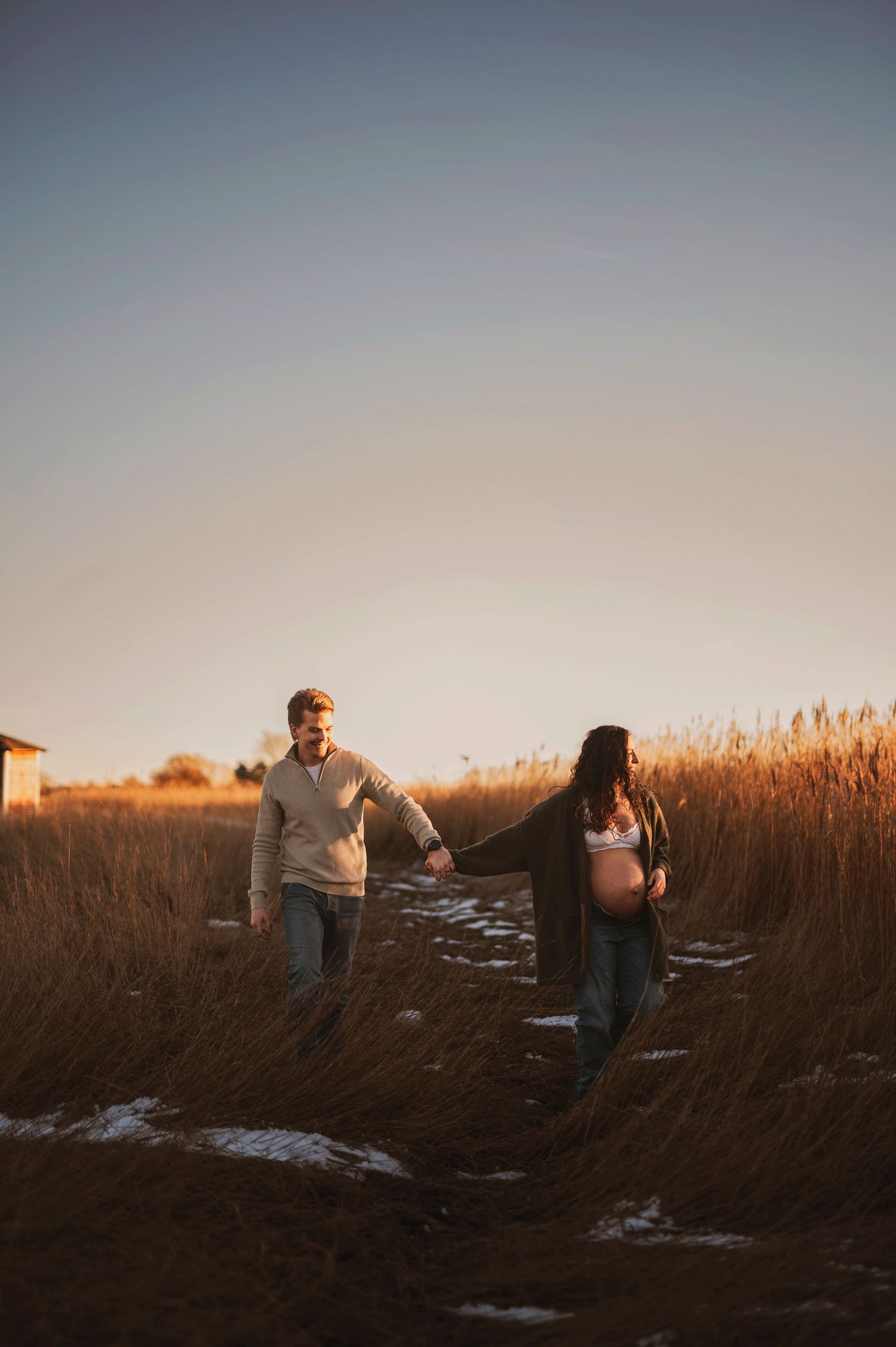 Couple walking hand in hand through a snowy field at sunset surrounded by tall grass.