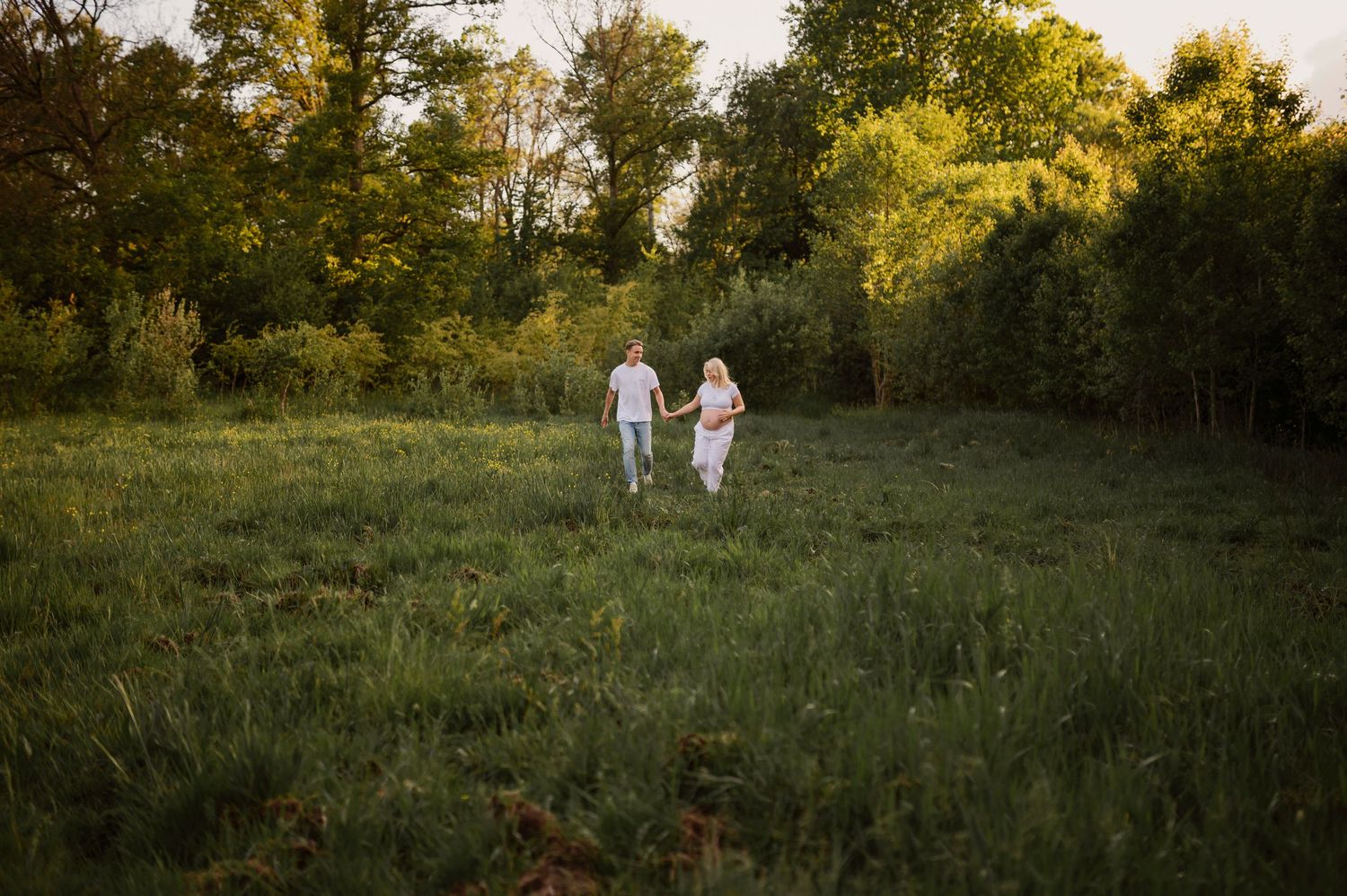 A couple in white clothing walks together through a sunlit meadow surrounded by green trees at golden hour.